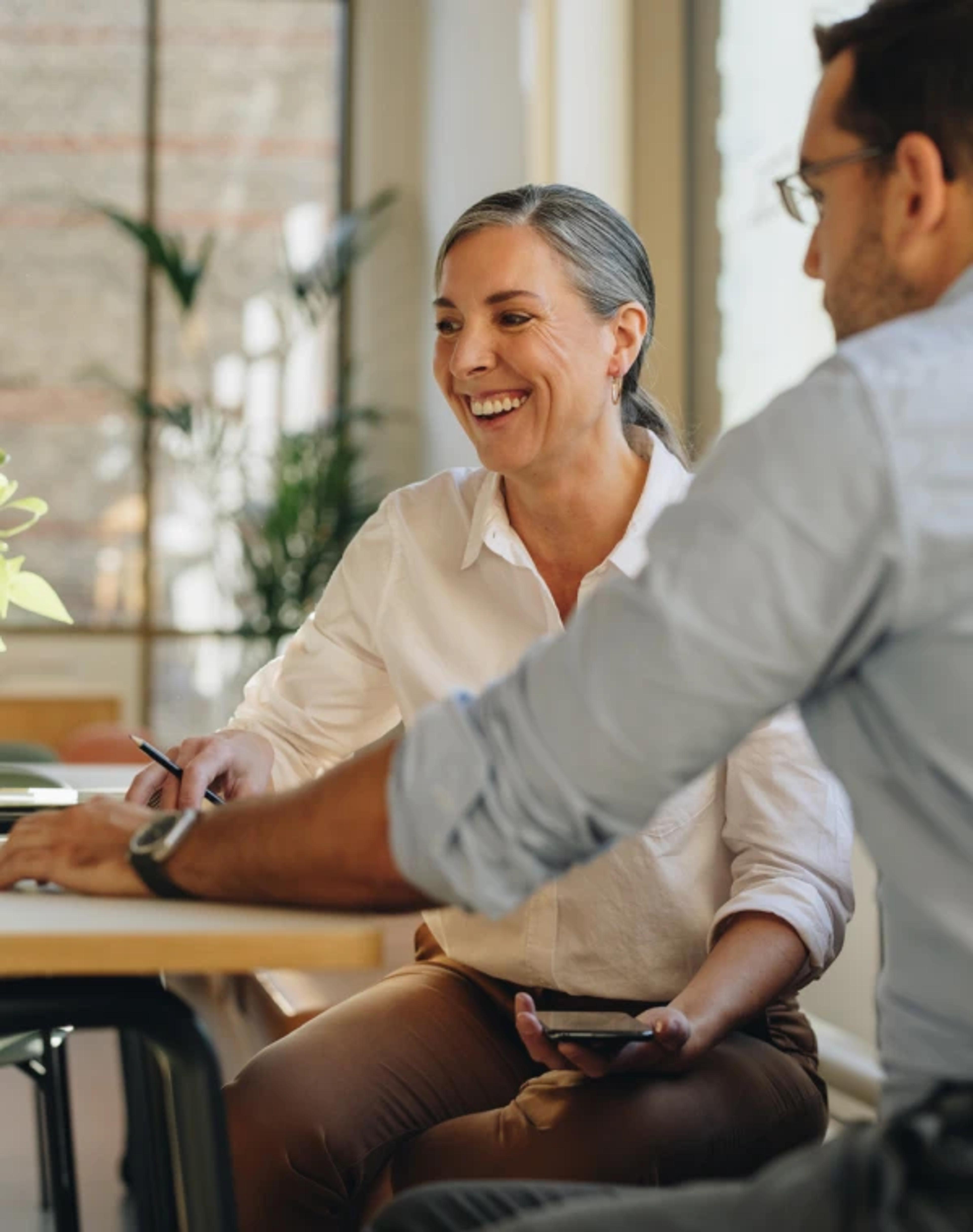 A smiling woman with grey hair tied back in a low ponytail sitting at a bright office table, holding a pen and a smartphone while talking to a colleague wearing a light blue shirt seen in the foreground.