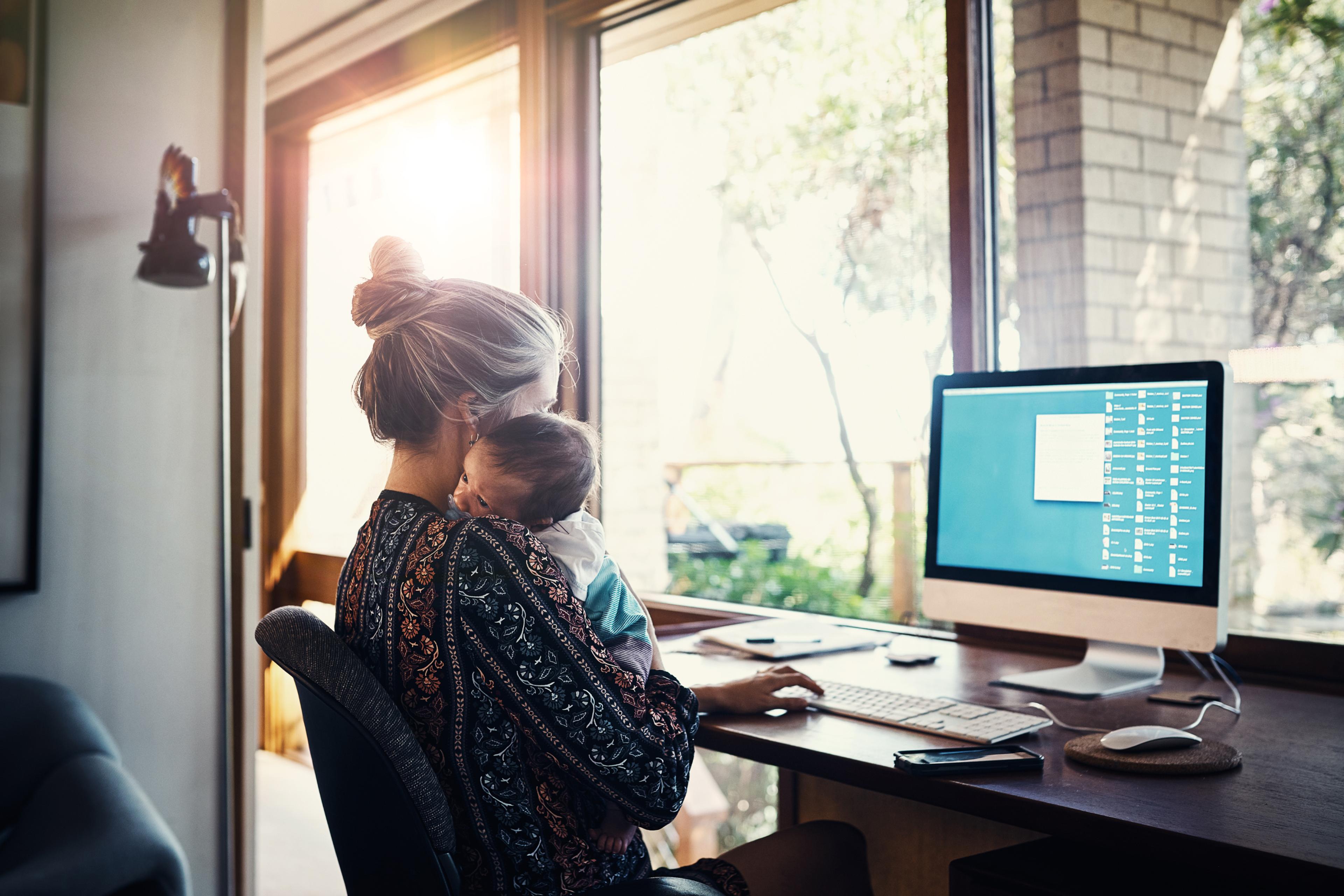 A mother holds her baby on her shoulder while working on a desktop computer.