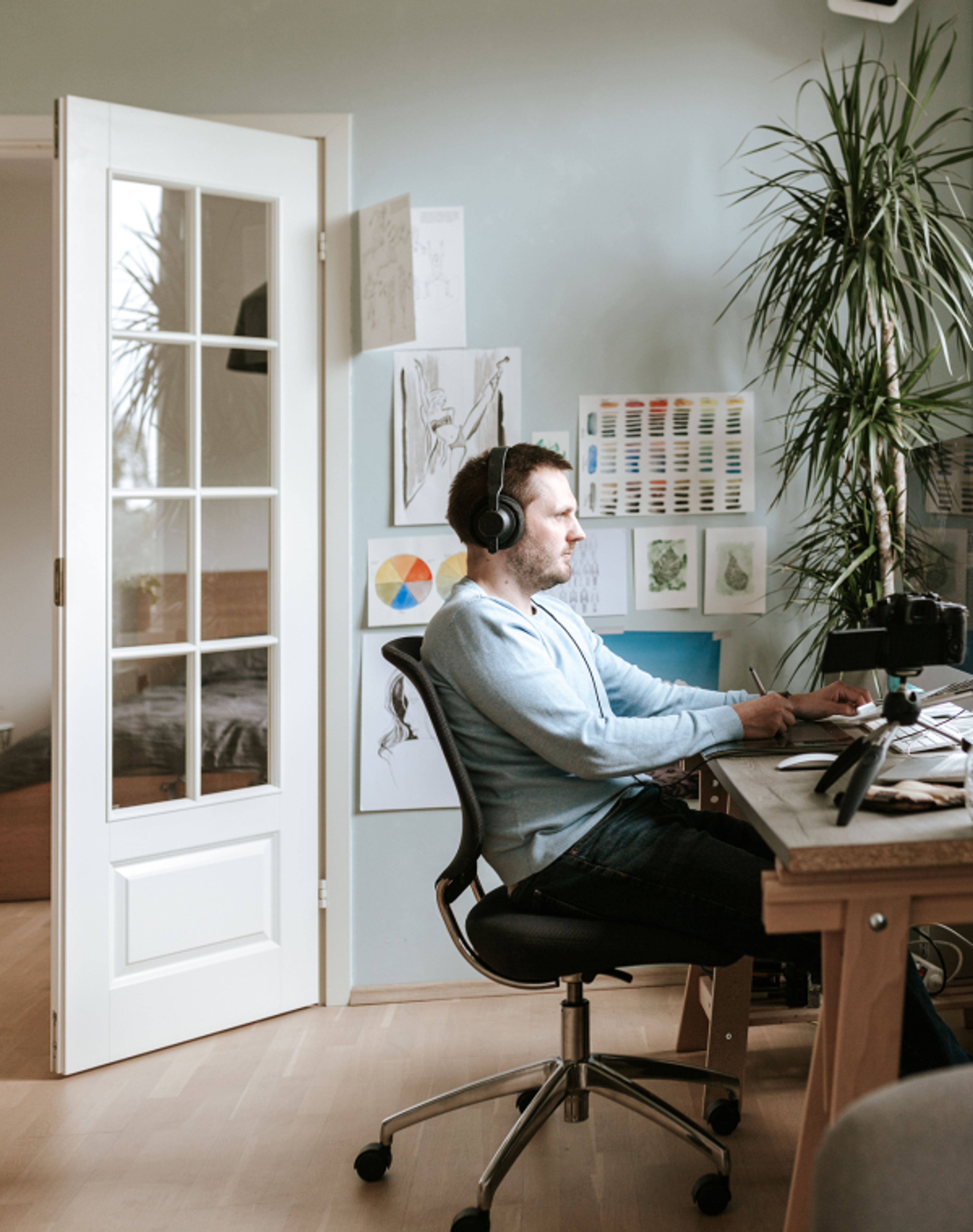 A professional woman with shoulder-length brown hair, wearing glasses, a white blouse, and wireless earbuds, sits at a wooden desk focusing on her laptop while taking notes on paper with a pen in her right hand.