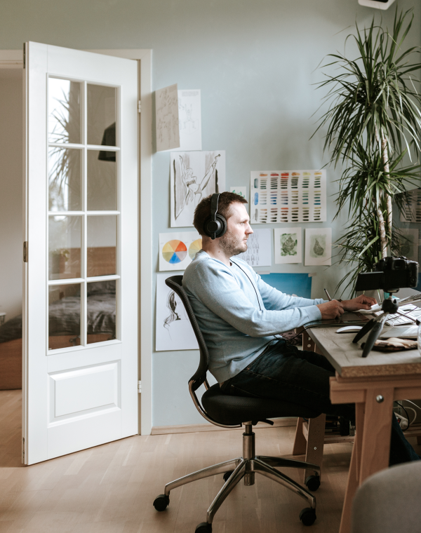 A professional woman with shoulder-length brown hair, wearing glasses, a white blouse, and wireless earbuds, sits at a wooden desk focusing on her laptop while taking notes on paper with a pen in her right hand.