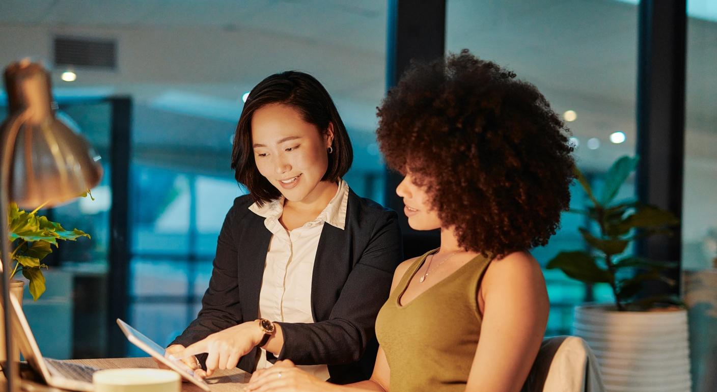 Two women sitting together at an office desk while one points at a tablet screen.