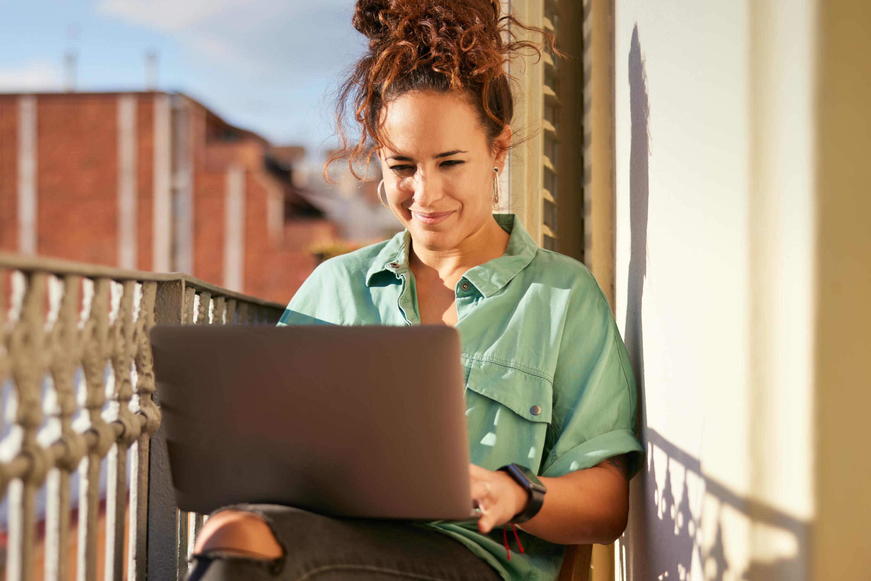 Young woman with curly hair working on a laptop on a sunny balcony.