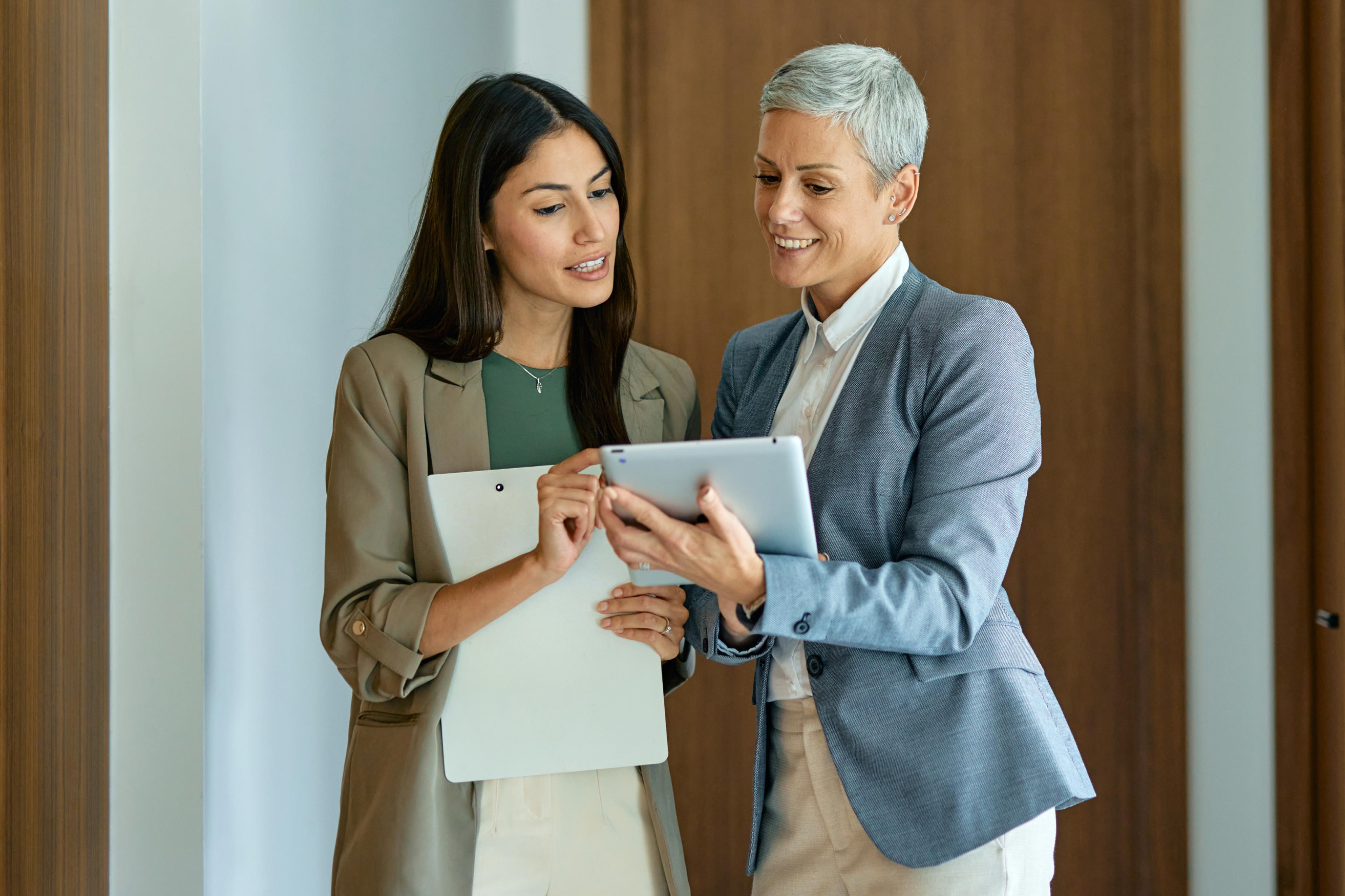 Two women in business attire look at a tablet together.