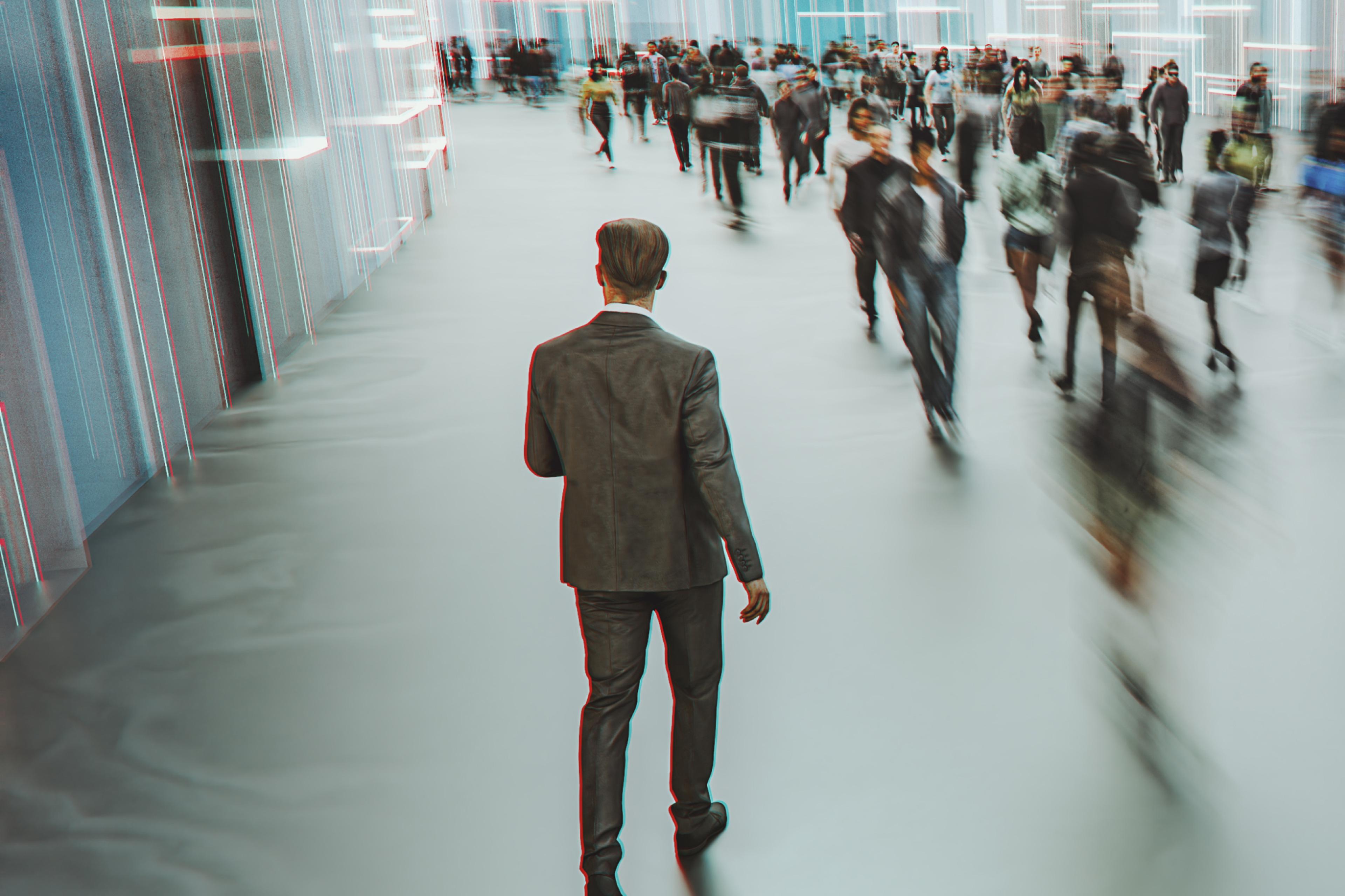 A man in a suit walks away from the viewer, past a motion-blurred crowd in a bright, modern hallway.