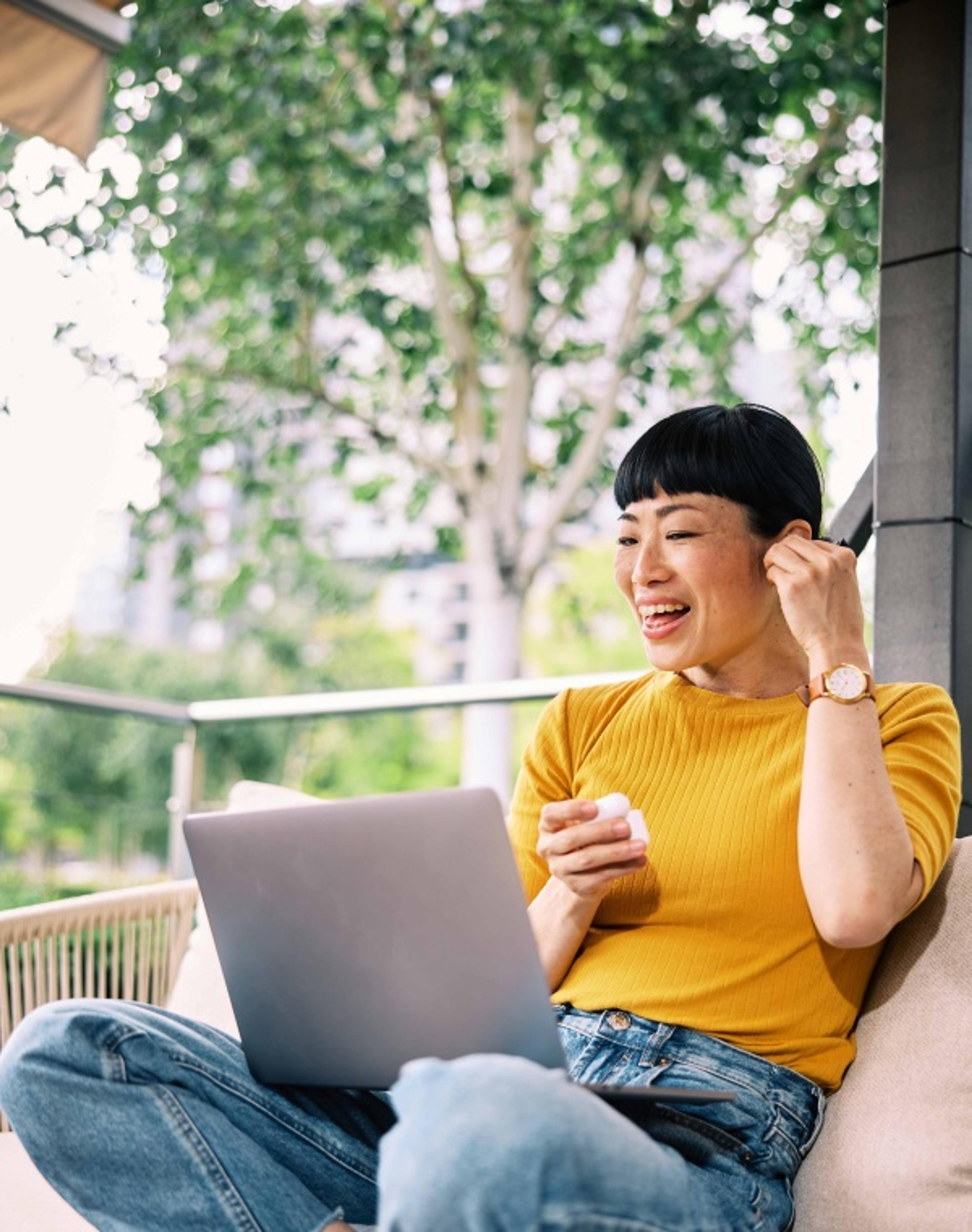 A woman in a yellow shirt sitting cross-legged with a laptop outdoors while adjusting an earbud.