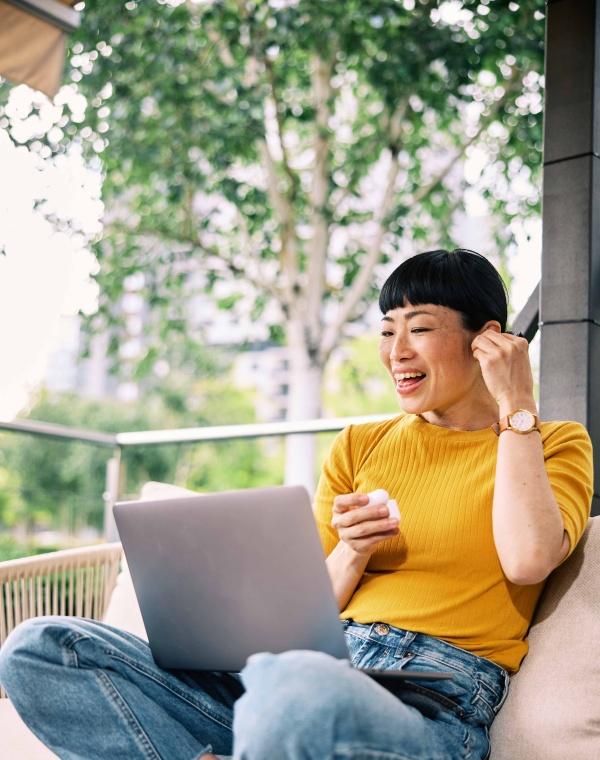 A woman in a yellow shirt sitting cross-legged with a laptop outdoors while adjusting an earbud.