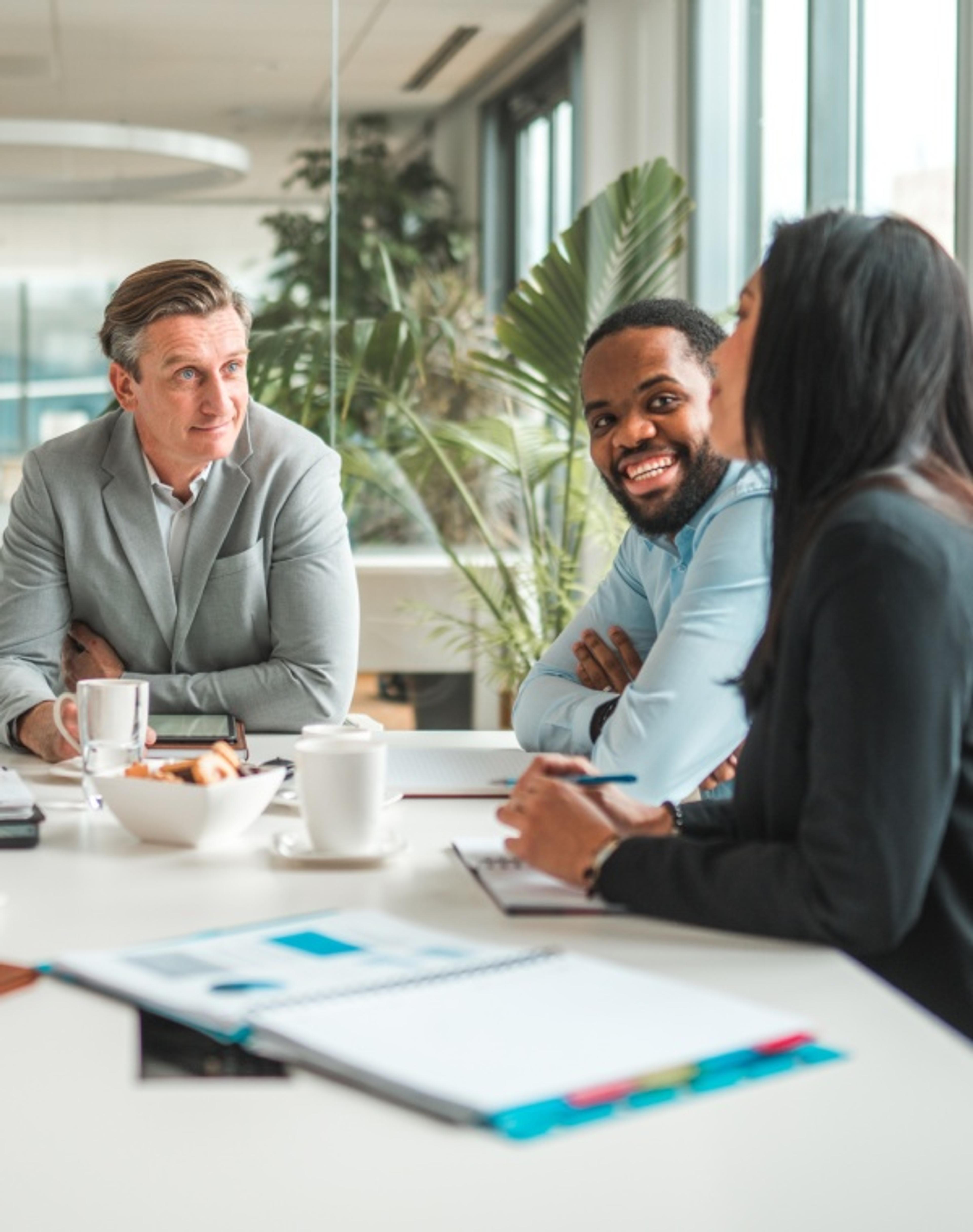 Three colleagues sitting around a conference table engaged in a meeting, with coffee cups, notebooks, and documents spread out.