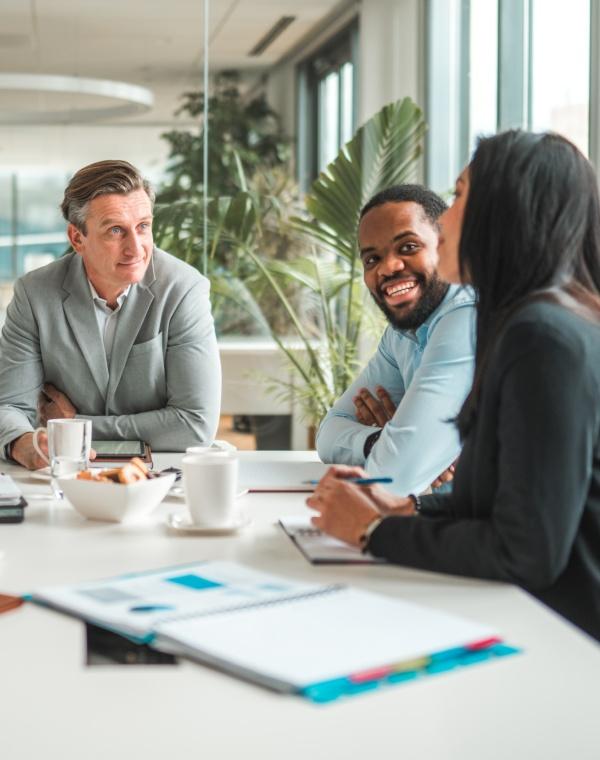 Three colleagues sitting around a conference table engaged in a meeting, with coffee cups, notebooks, and documents spread out.