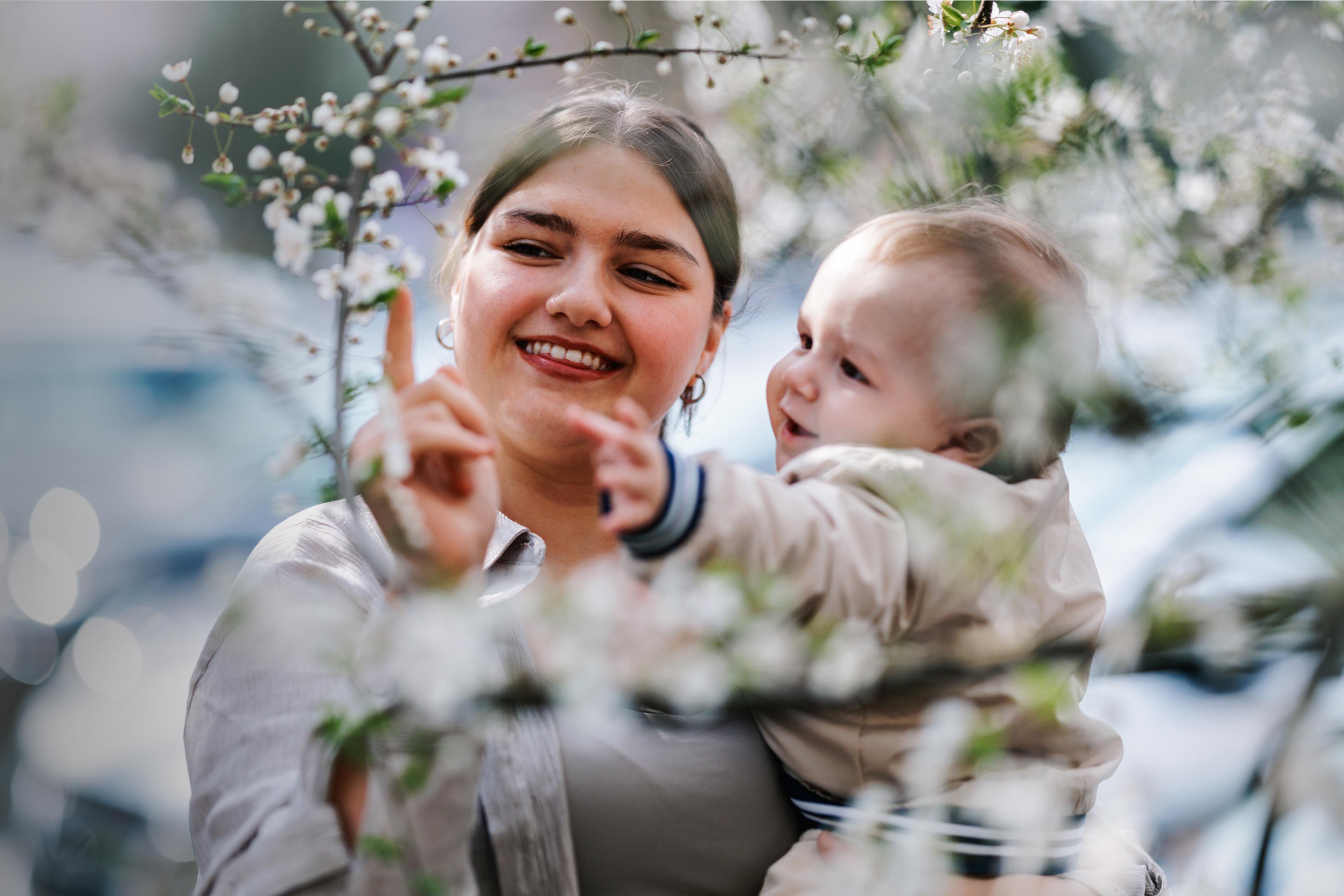 A smiling woman holds a baby, pointing at white blossoms while the baby also reaches out.