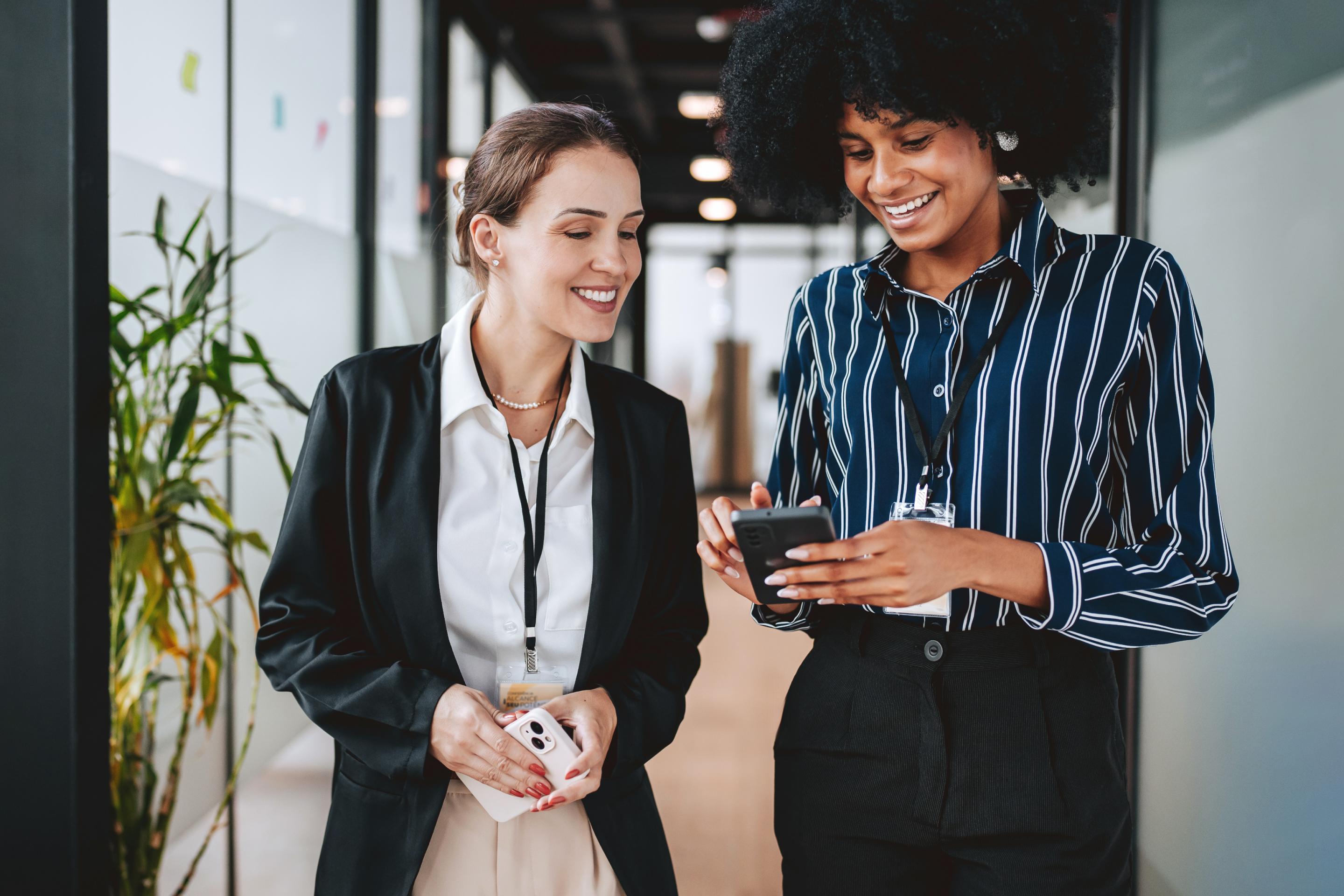 Two professional women smiling and looking at a smartphone in an office hallway.
