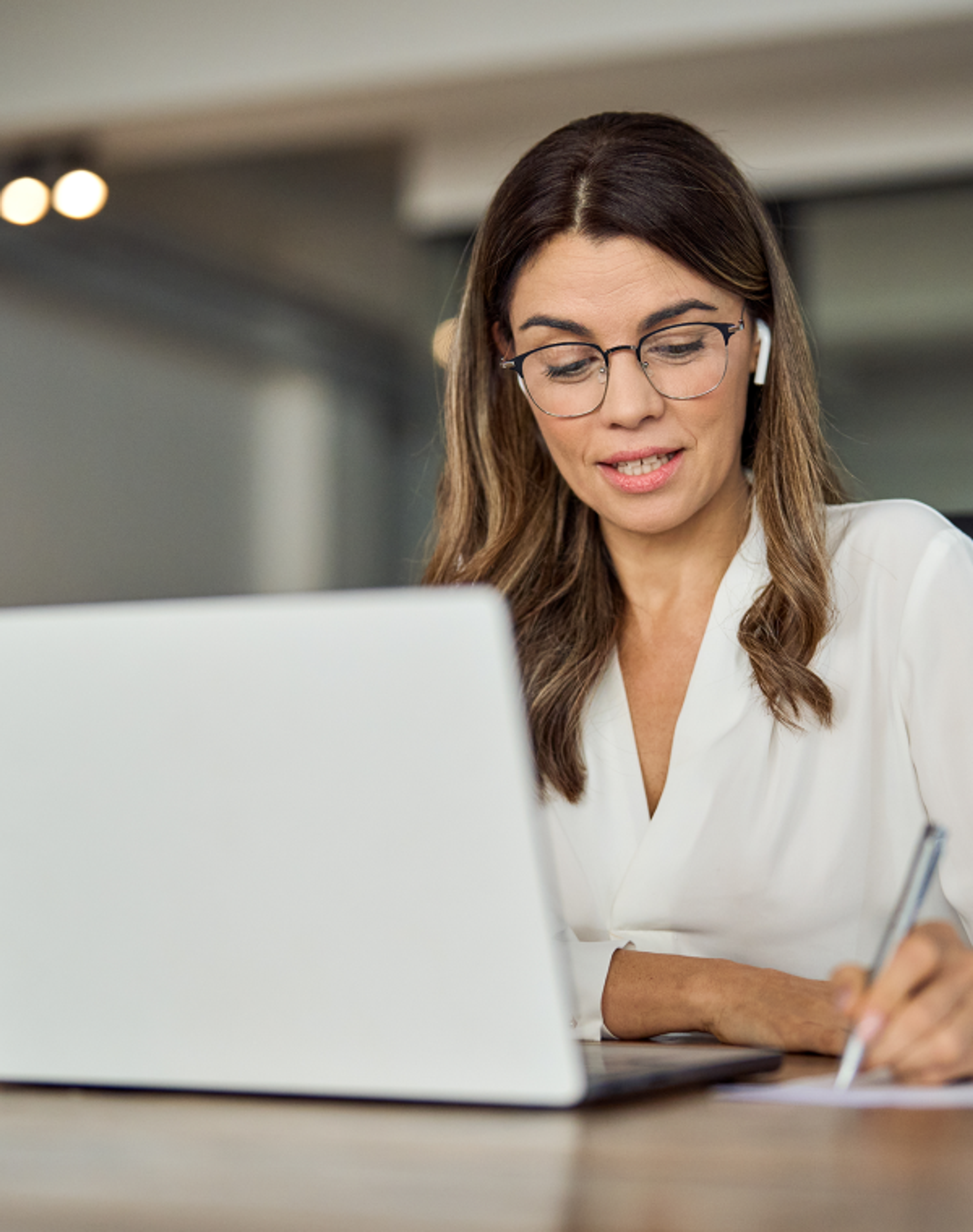 A professional woman with shoulder-length brown hair, wearing glasses, a white blouse, and wireless earbuds, sits at a wooden desk focusing on her laptop while taking notes on paper with a pen in her right hand.