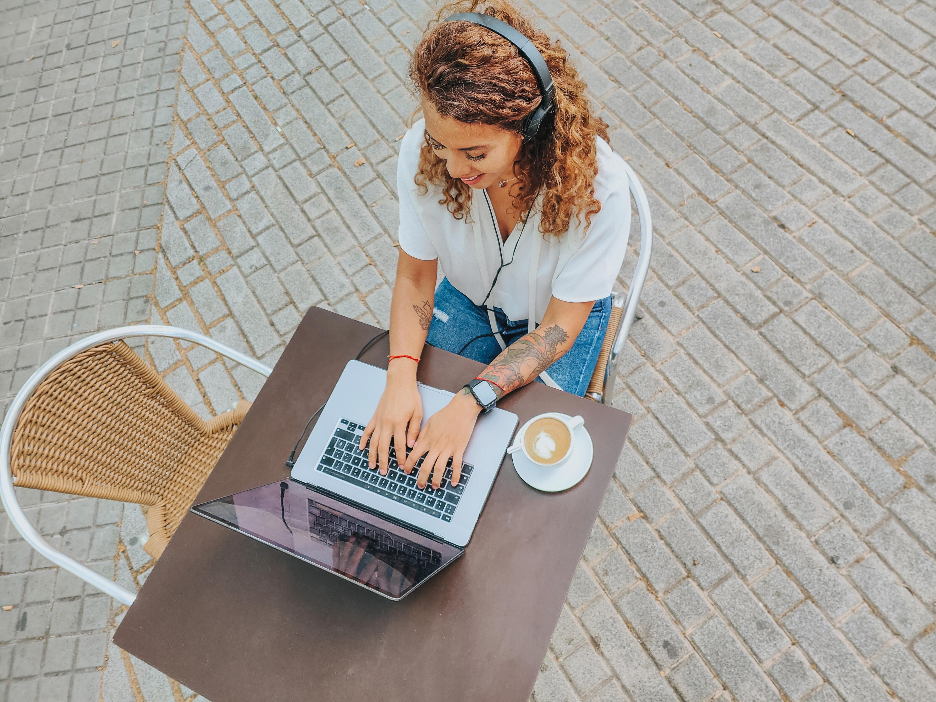 Birds-eye-view of remote worker outdoors, sitting at cafe table with coffee, typing on laptop.