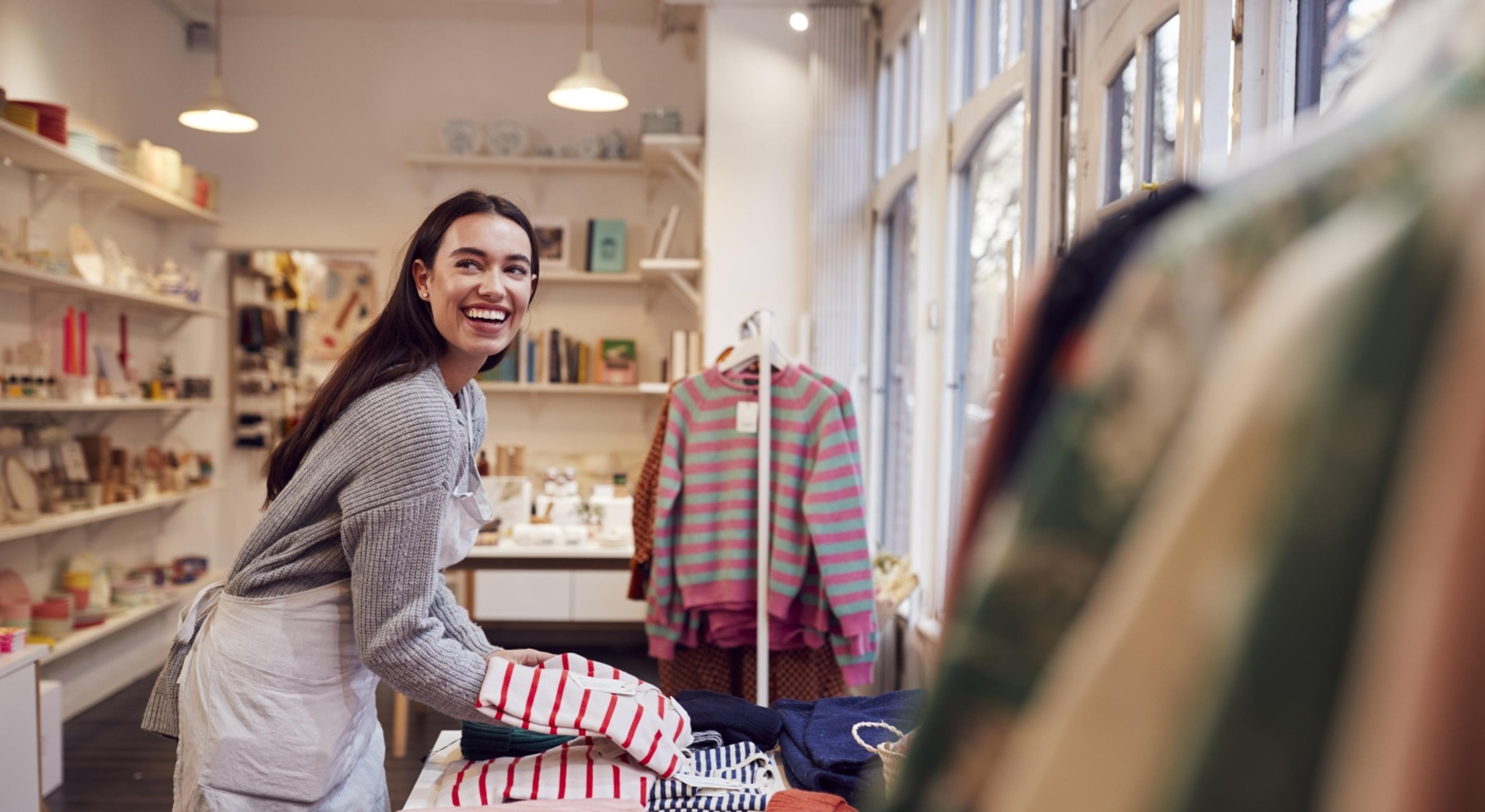 A smiling boutique worker in a grey sweater and white apron organizing folded clothing on a store display table.