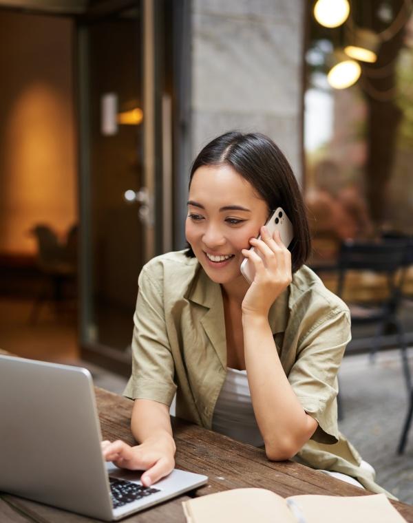 A smiling woman talking on the phone while using a laptop at an outdoor table.