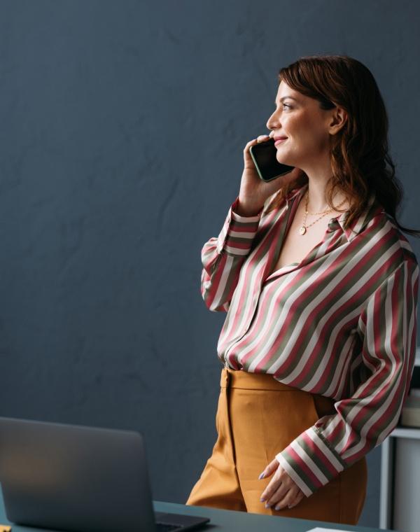 A smiling woman talks on a phone near a desk laptop.