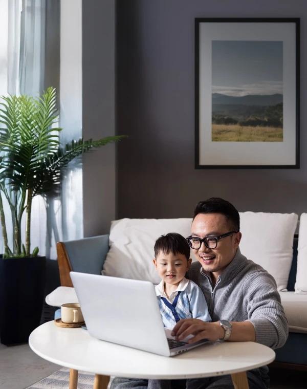 A smiling father sitting on the floor using a laptop while his young son sits beside him in a living room.