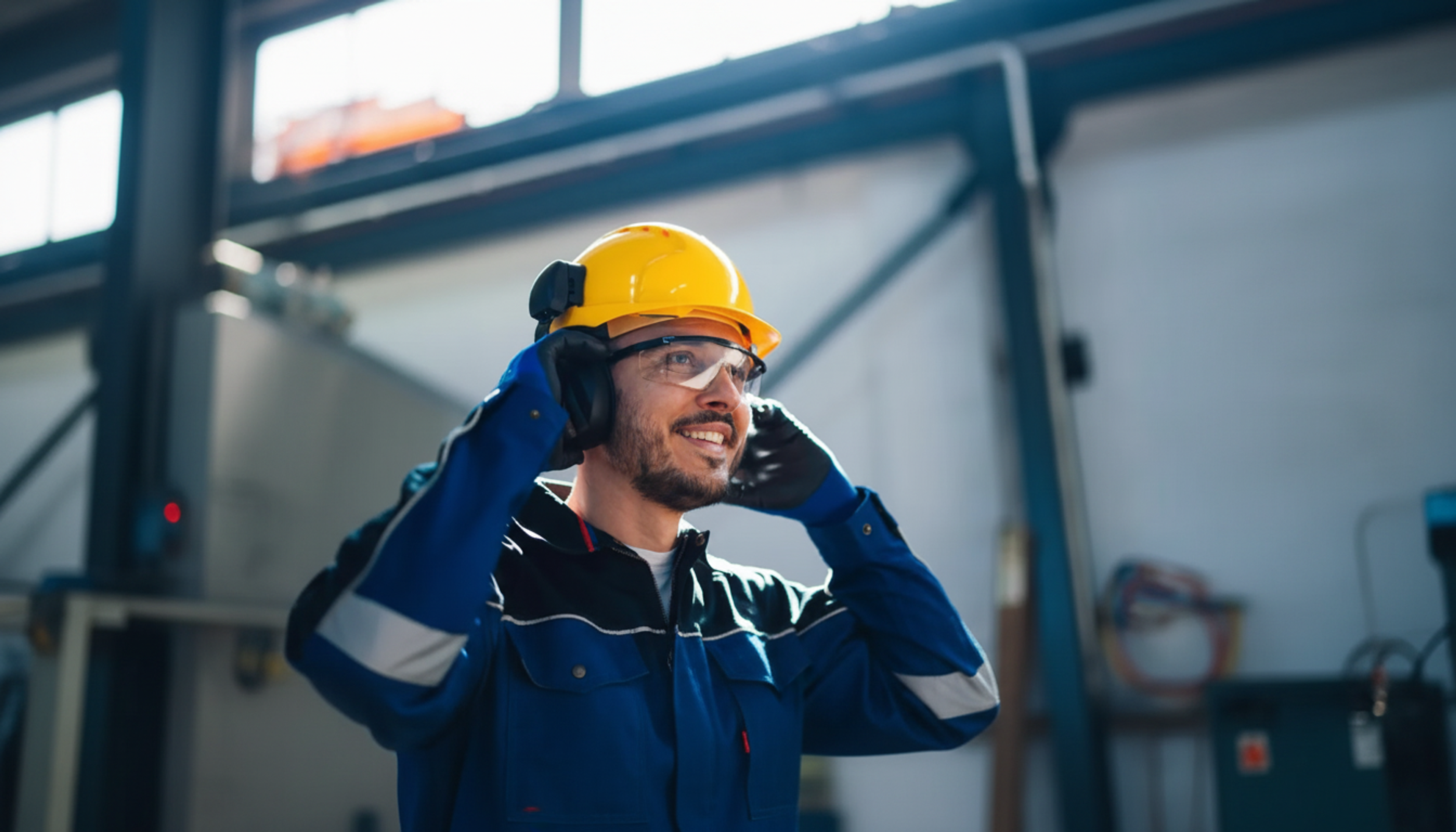 A smiling industrial worker putting on ear protection while wearing a yellow hard hat and safety glasses.