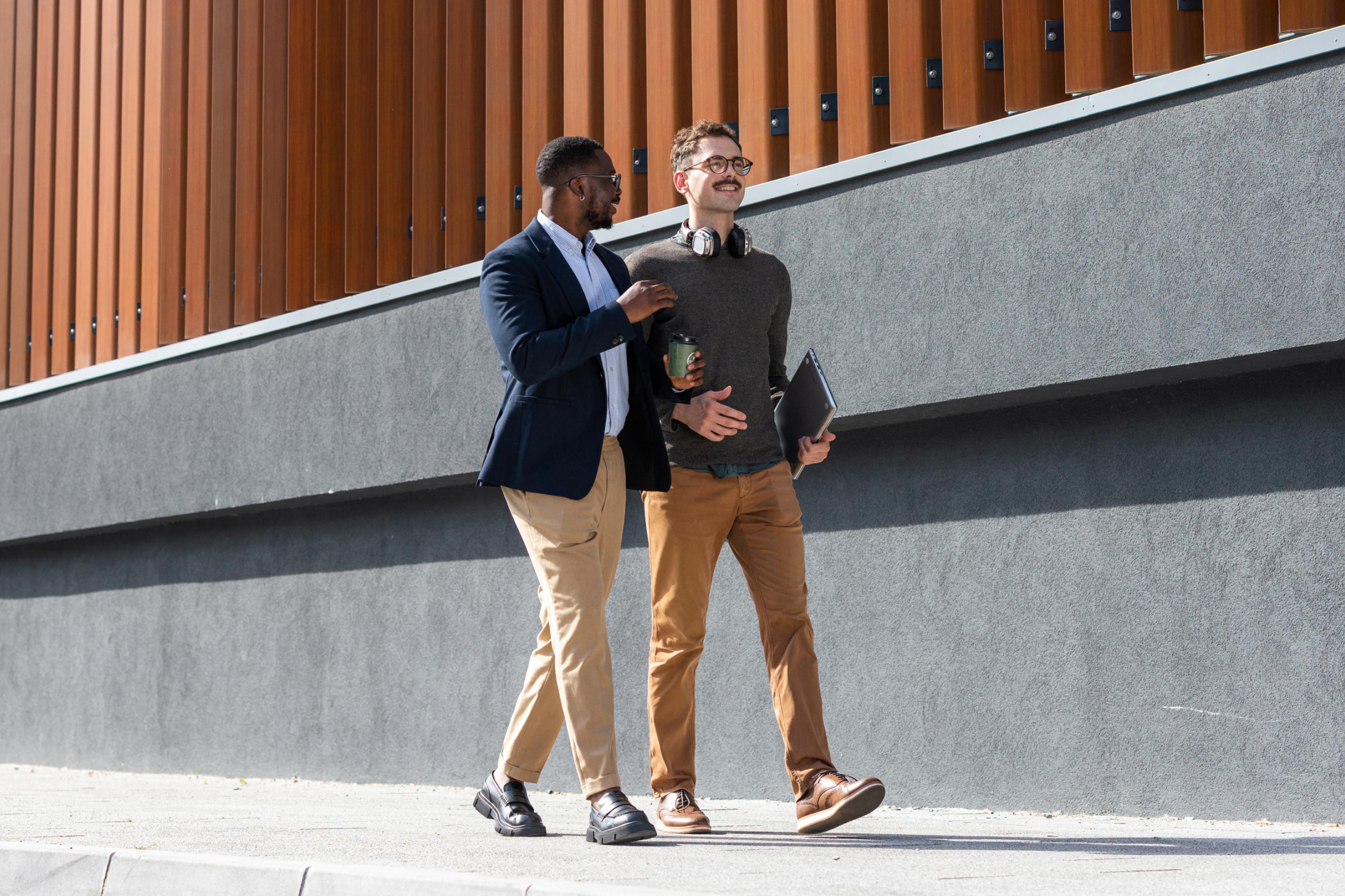 Two smiling men walk together on a sidewalk, one with an arm on the other's shoulder. One holds a laptop and coffee.