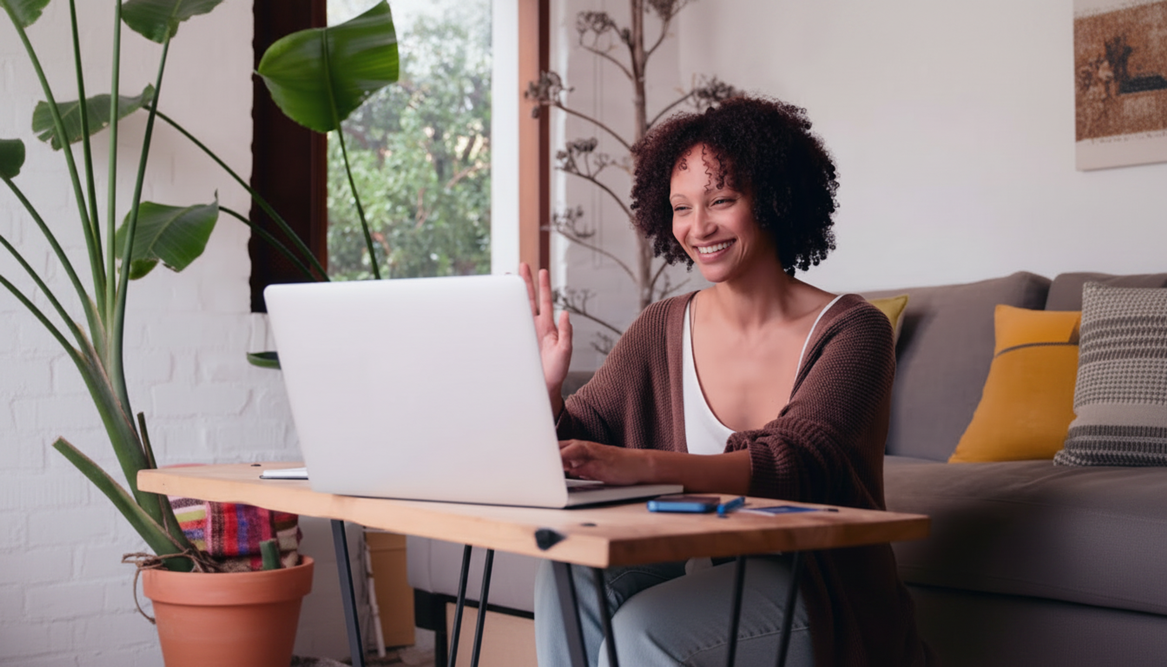 A smiling woman waving at her laptop during a video call at a desk.