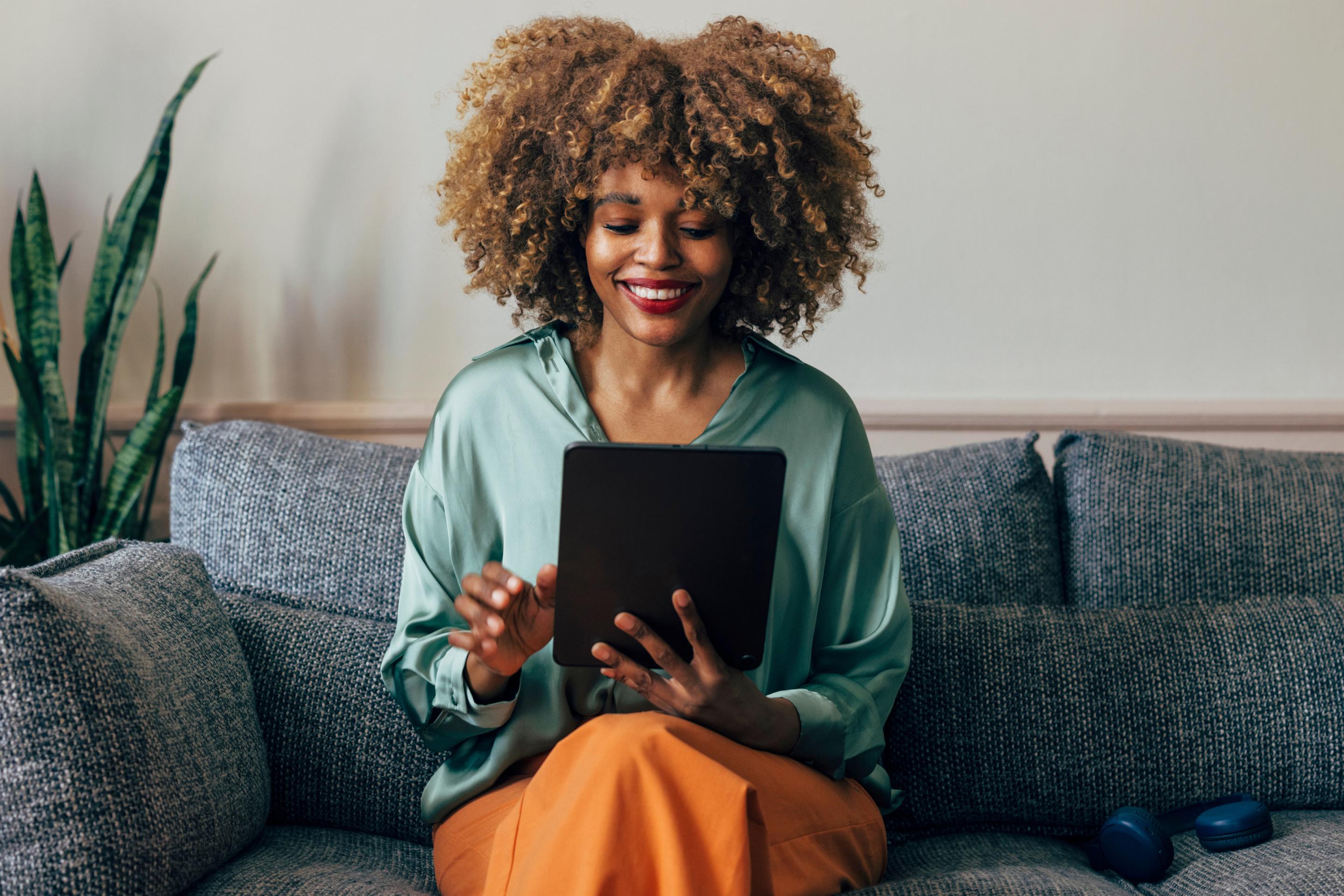 Smiling Black woman with curly hair sitting on a sofa and using a tablet.