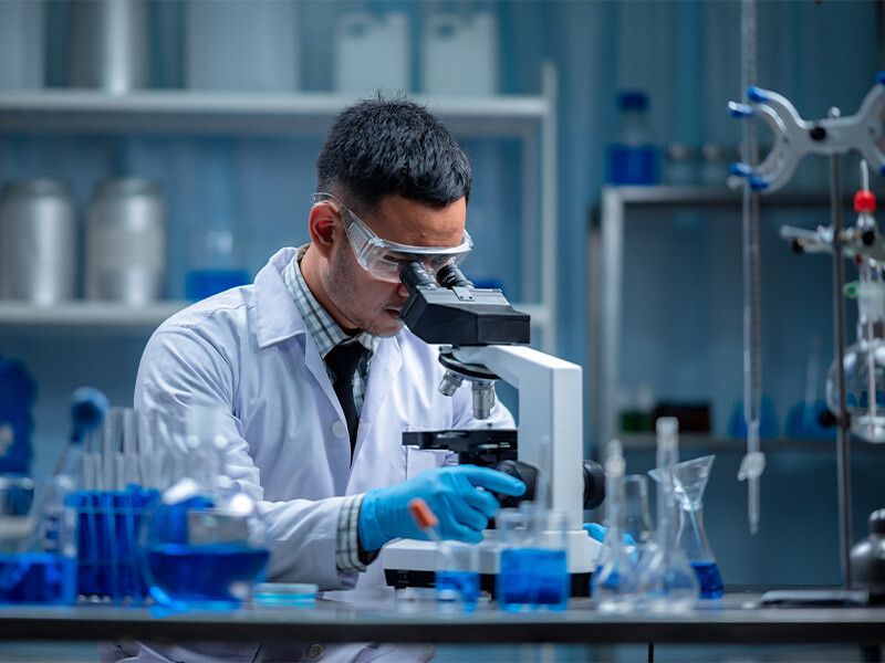 A scientist in a lab coat, safety glasses and gloves looking through a microscope at a bench of glassware