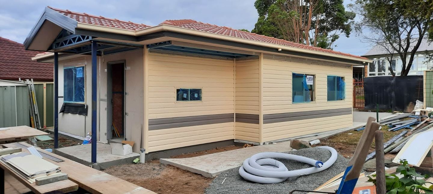 The dwelling clad in weatherboard-profile sheeting with contrasting bands, windows glazed and the tiled roof complete