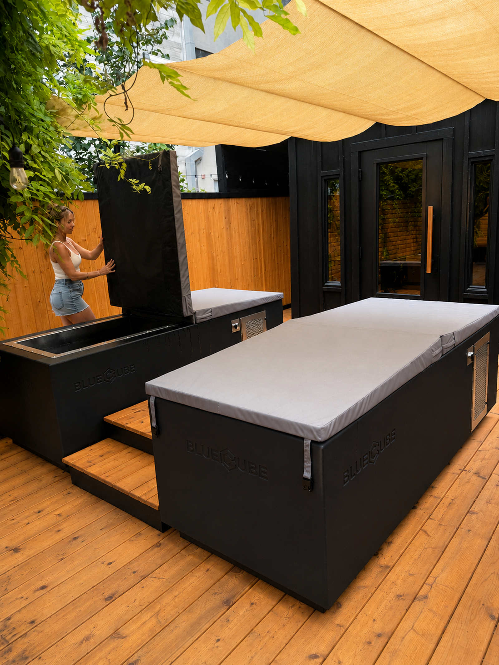 A woman opens a black BlueCube ice bath on an outdoor wooden patio with another covered BlueCube next to it.