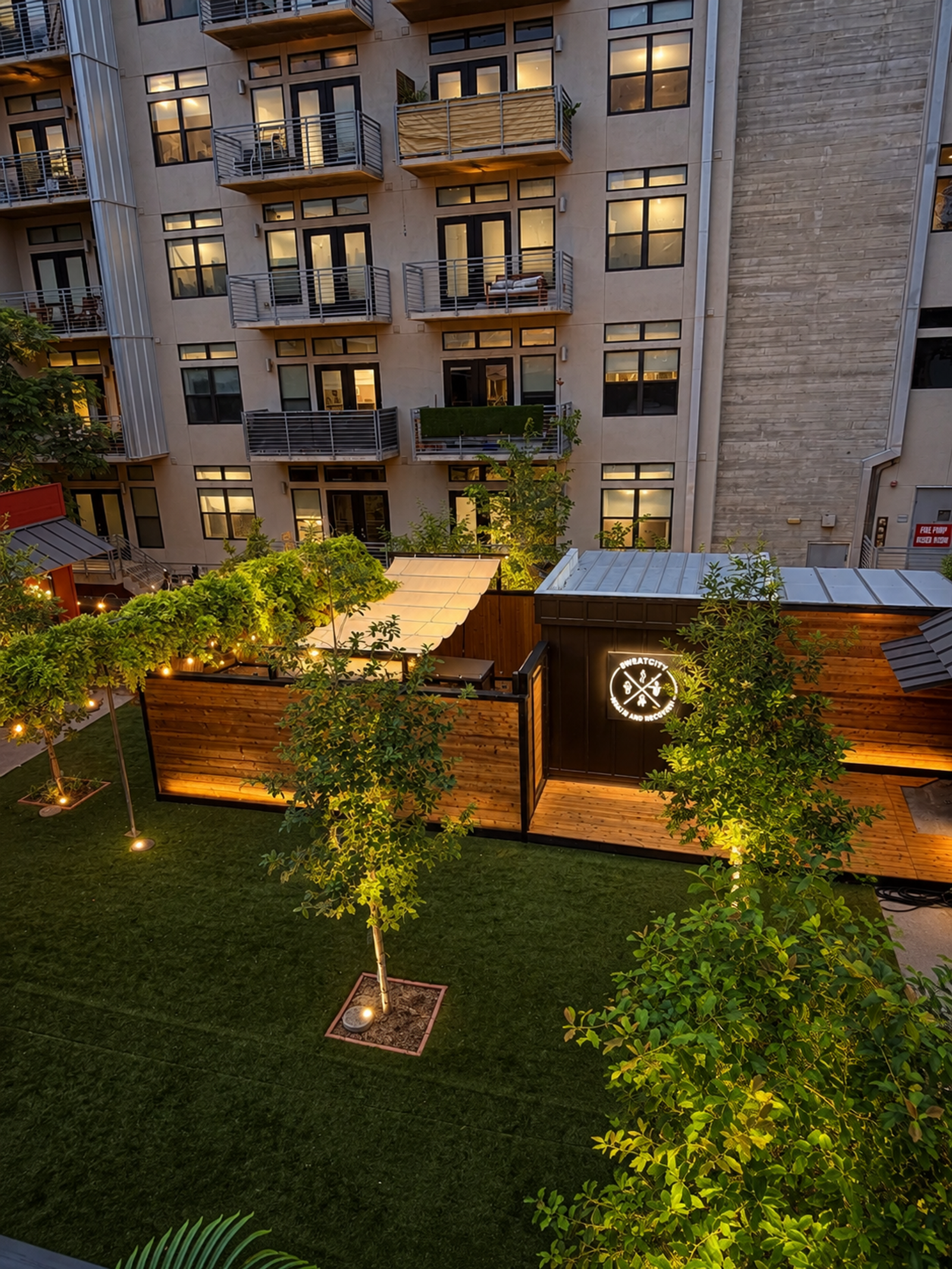 Overhead view of an outdoor axe throwing venue with a lawn, illuminated trees, string lights, and a modern building in the background at dusk.