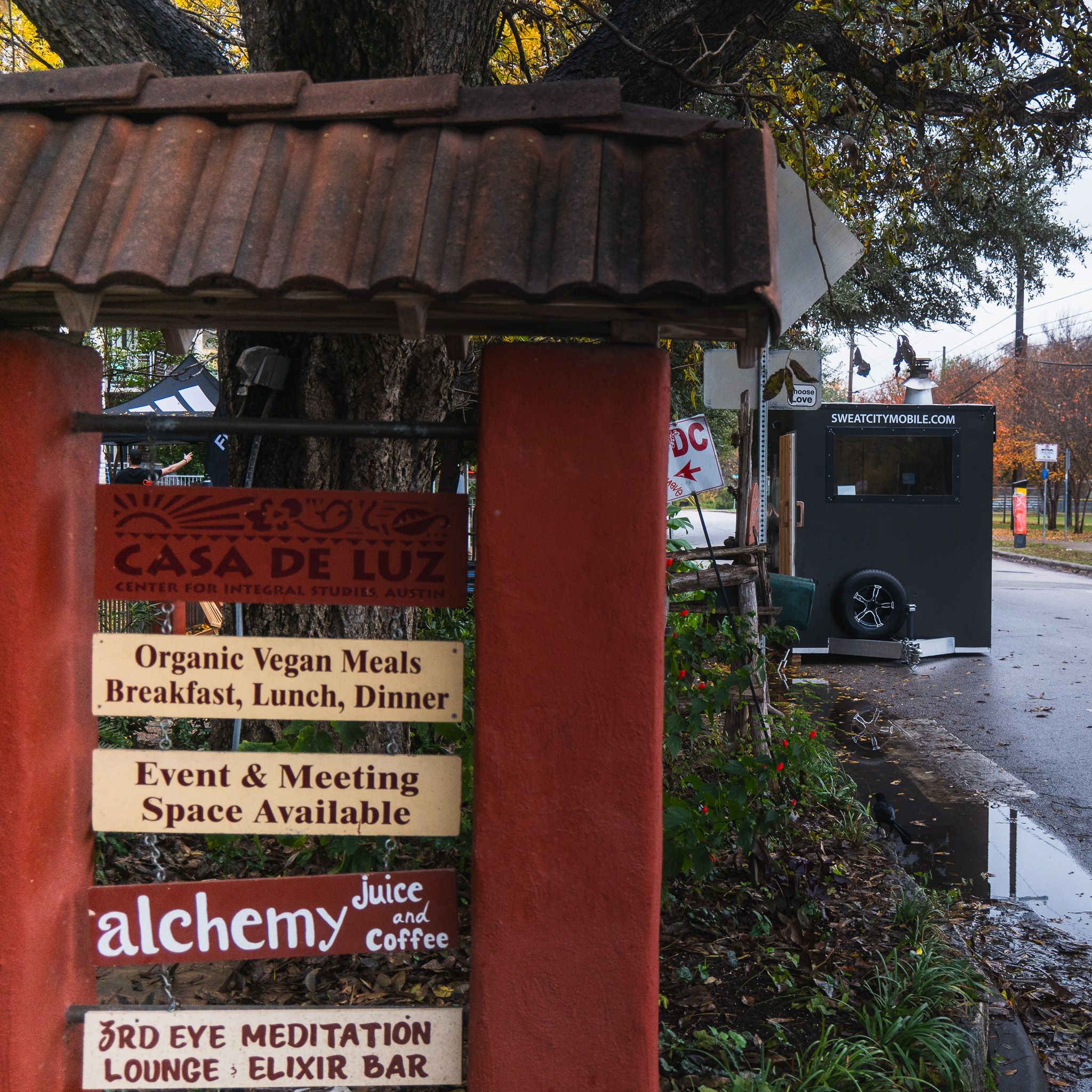 A red multi-panel sign for "Casa de Luz" and related businesses stands next to a street with a black mobile sauna trailer.