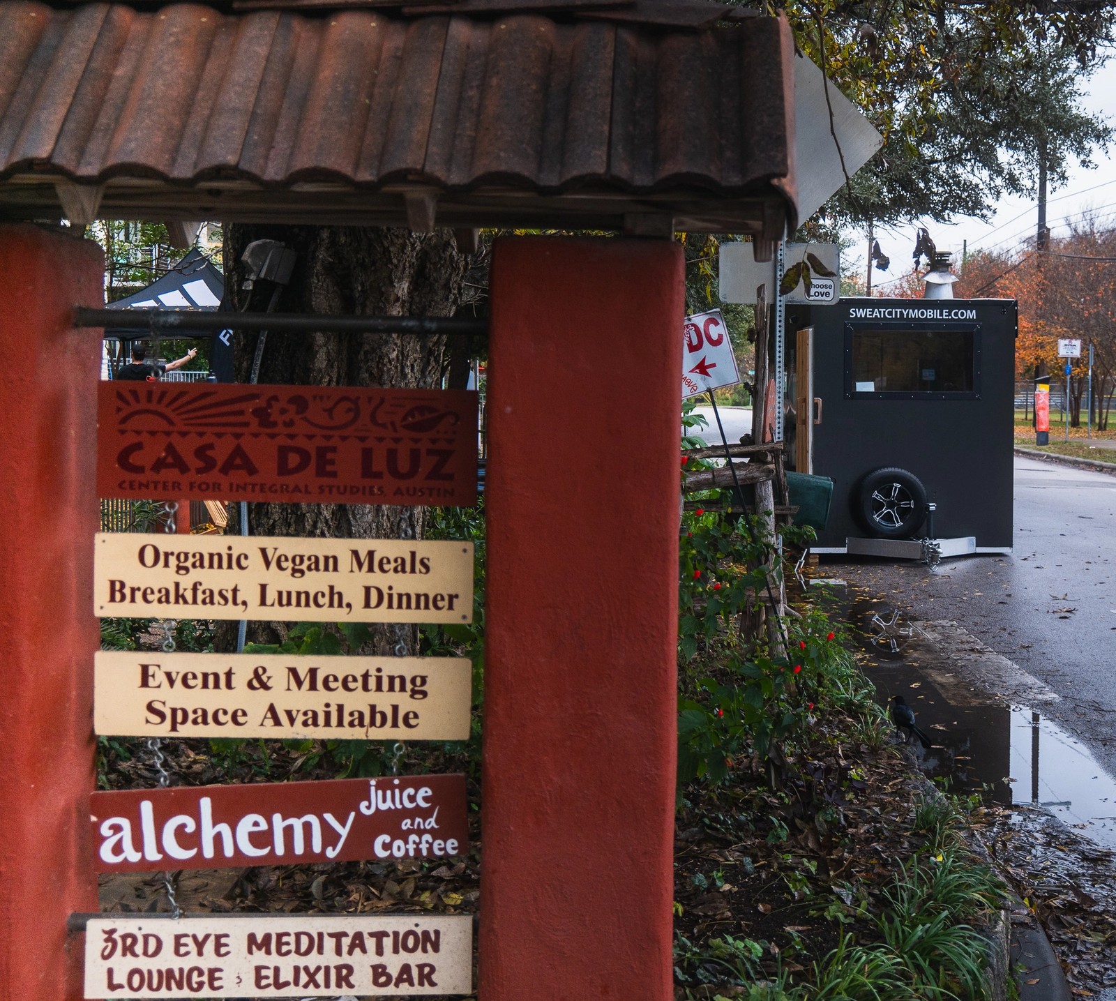 A red multi-panel sign for "Casa de Luz" and related businesses stands next to a street with a black mobile sauna trailer.