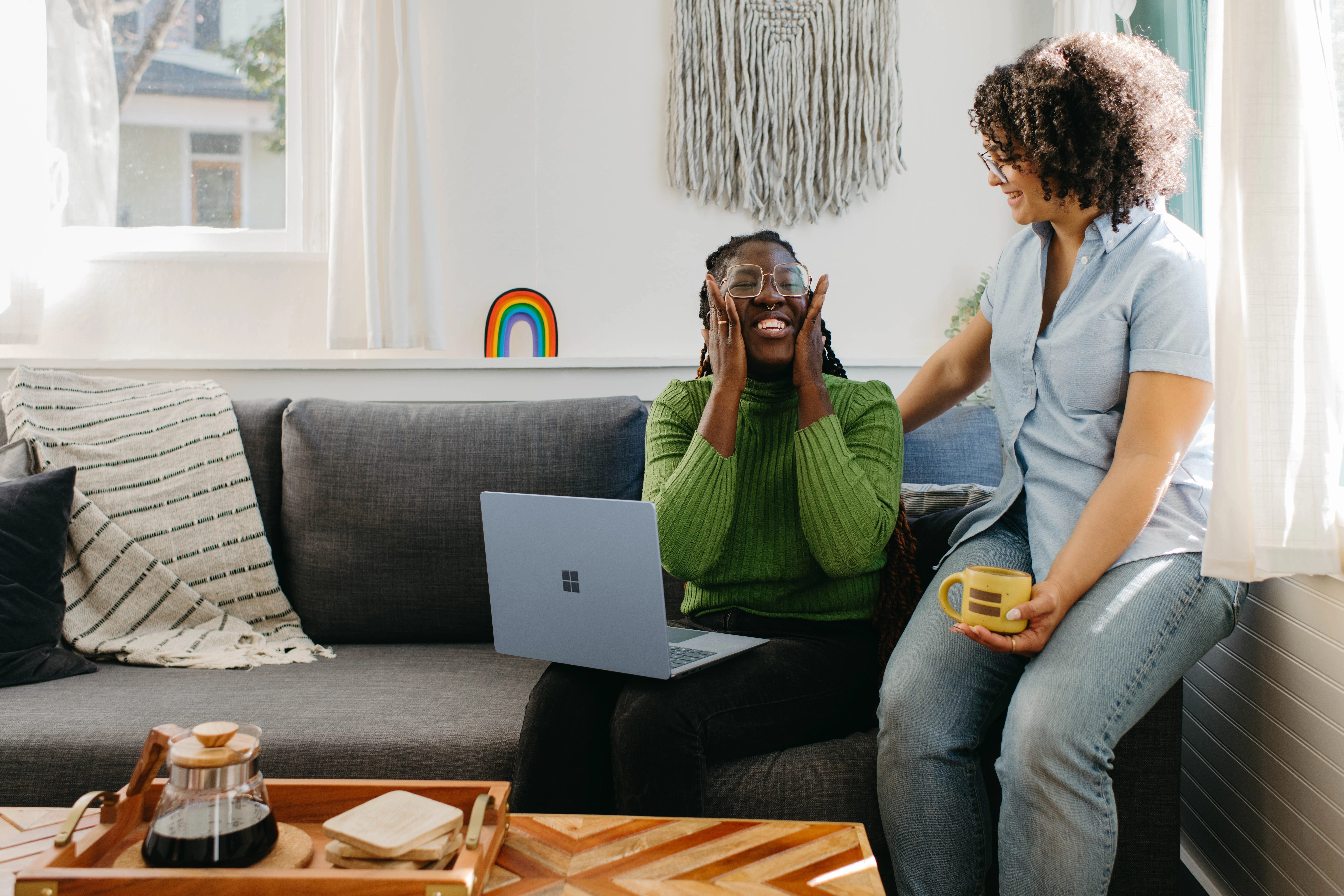 Two women are smiling, sitting on a grey sofa with a laptop, looking at the Privy app