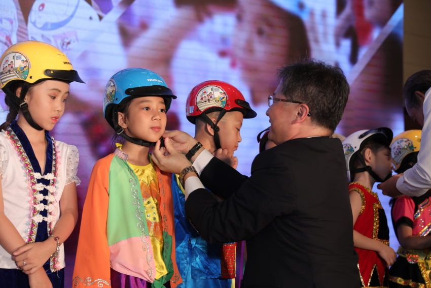 A stakeholder at the announcement ceremony assists a first grade student with putting on her new helmet.