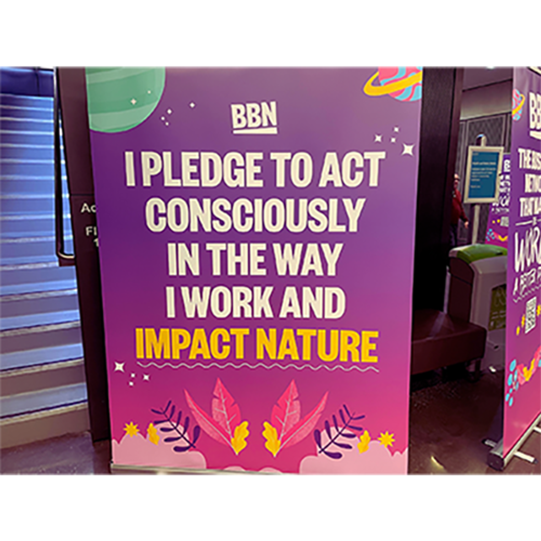 A multicoloured sign post which says in white and yellow coloured font, 'BBN I pledge to act consciously in the way I work and impact nature'. There is also illustrations of planets and nature on the poster. 