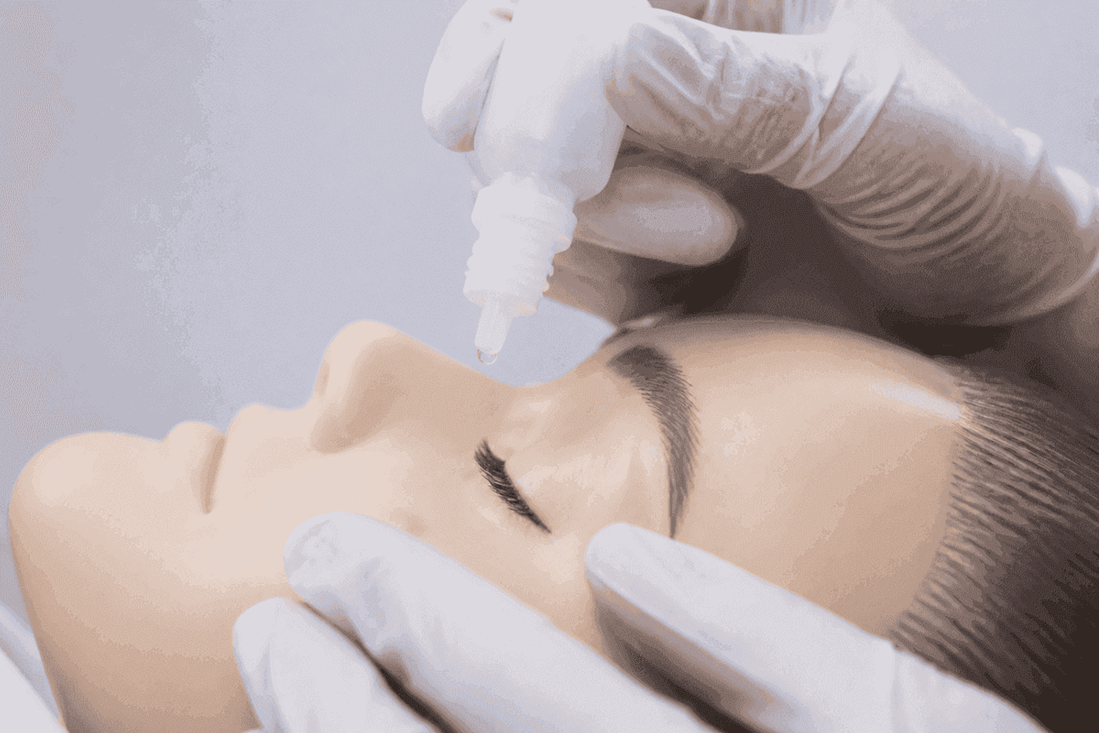 Gloved hands positioning an unmarked drop bottle above a closed eye for teaching technique.