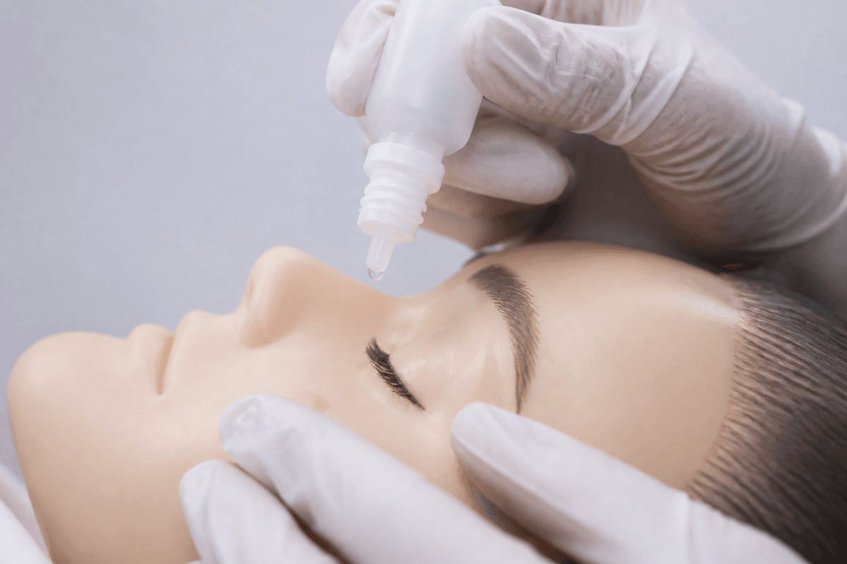 Gloved hands positioning an unmarked drop bottle above a closed eye for teaching technique.