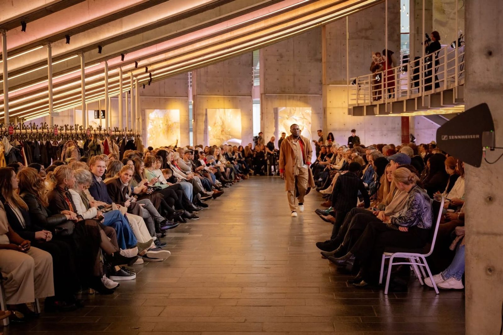 Défilé de mode dans une grande salle, mannequin en manteau beige marchant entre deux rangées de public — l'ambiance d'une fashion week nordique.