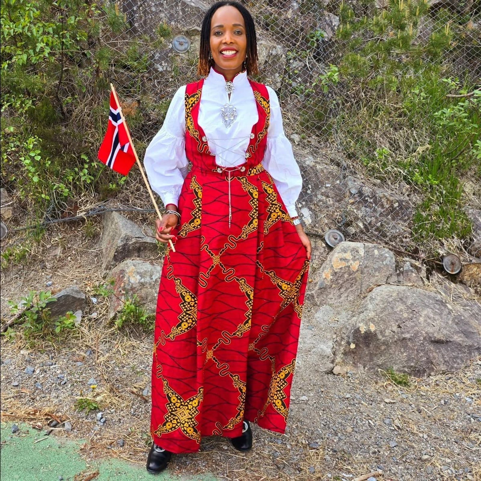 Femme portant un bunad norvégien réinterprété en tissu wax africain, drapeau norvégien à la main — symbole de la rencontre entre héritage africain et mode nordique.