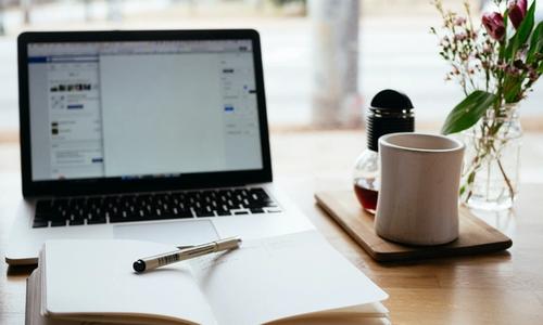 An open notebook on a table, with a pen sitting on top of the pages, with a laptop and a coffee mug in the background.