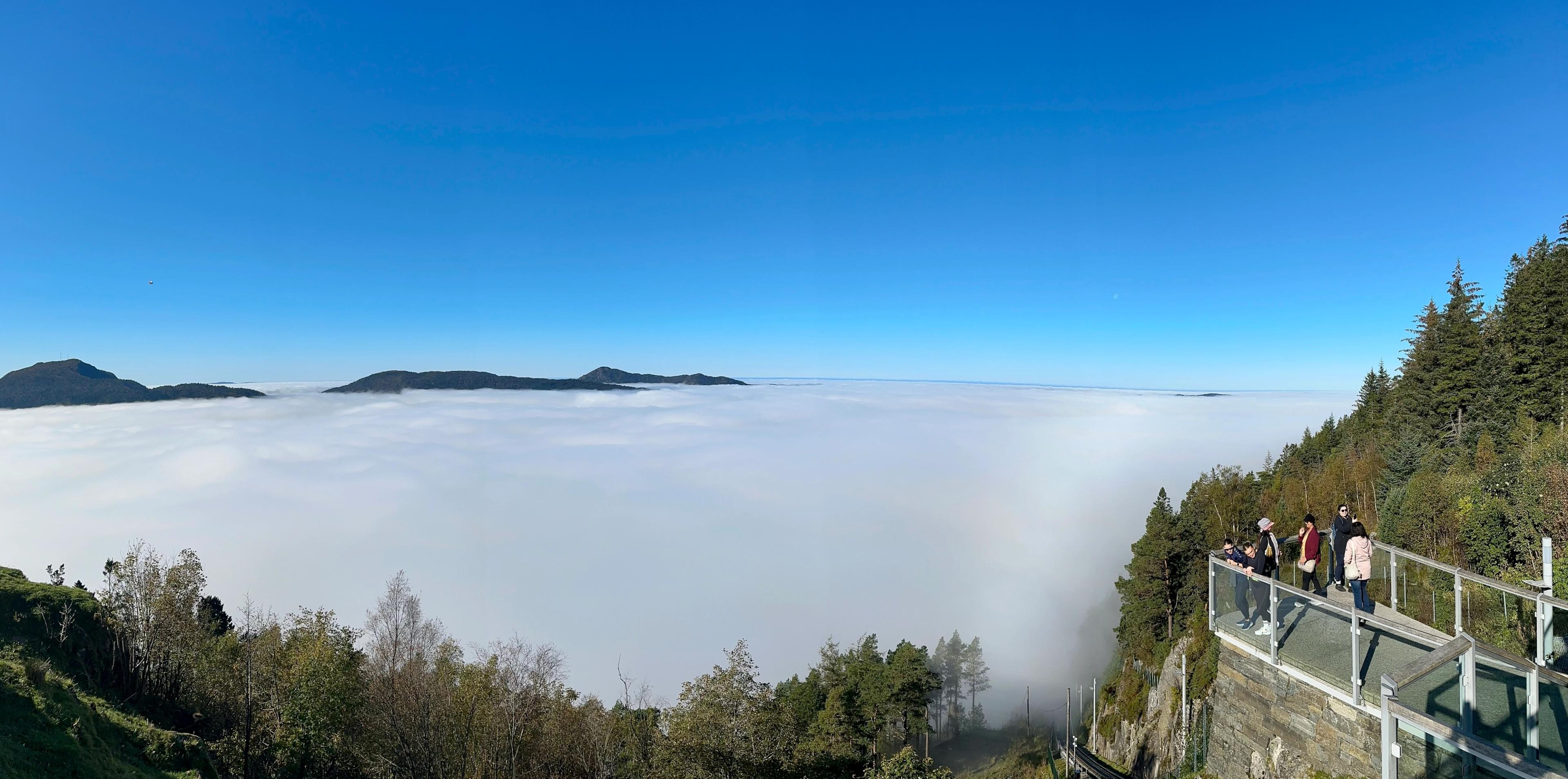 Panorama fra utsiktsplattformen på Fløyen over et tett skylag som dekker hele byen. Bare de omkringliggende fjelltoppene stikker opp av tåka, himmelen over er skyfri, og noen mennesker står ved glassrekkverket til høyre.