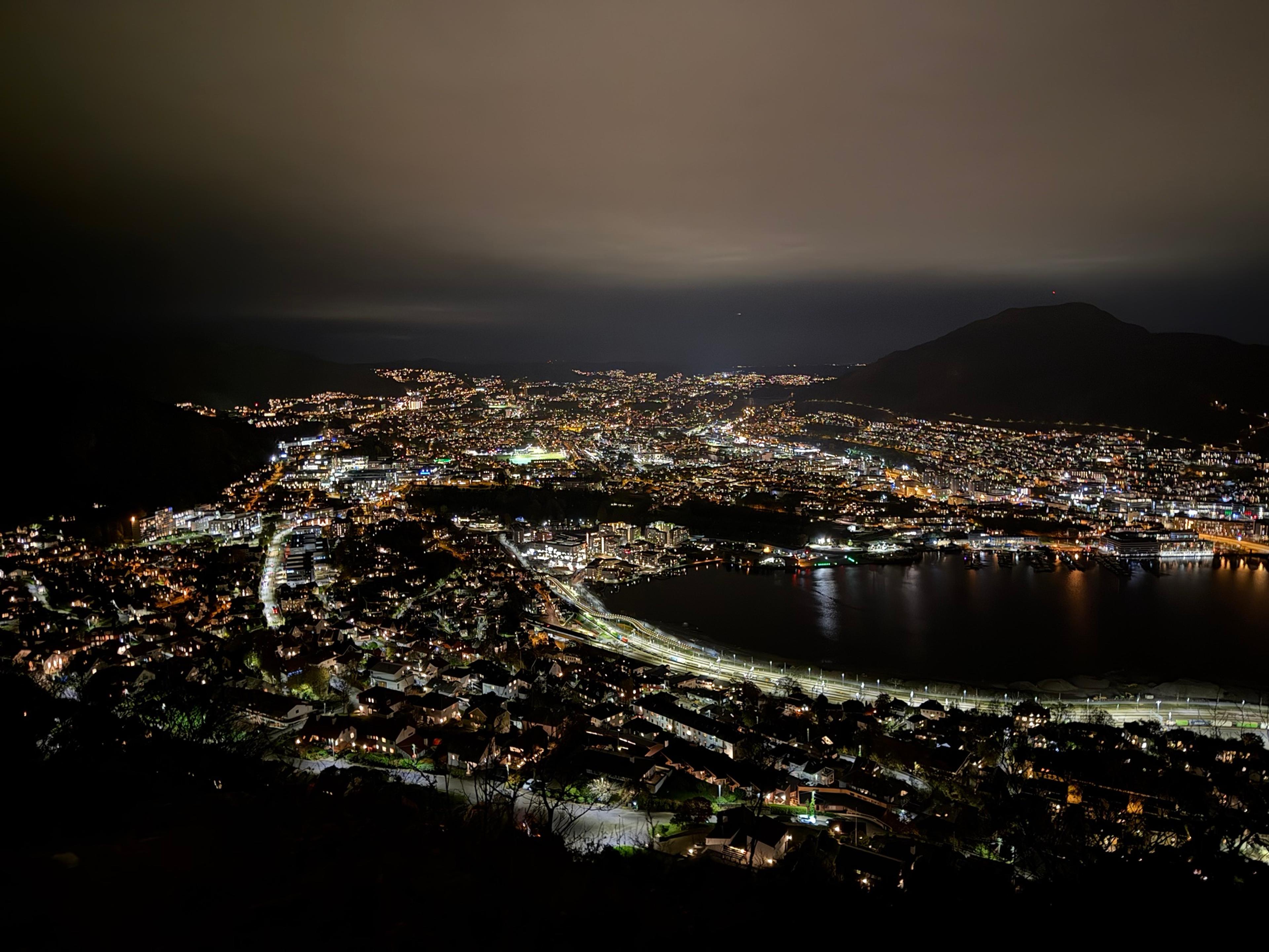 Bergen i kveldsmørke sett ovenfra, med et vann der jernbanen går i en opplyst bue langs bredden og et mørkt fjell til høyre.