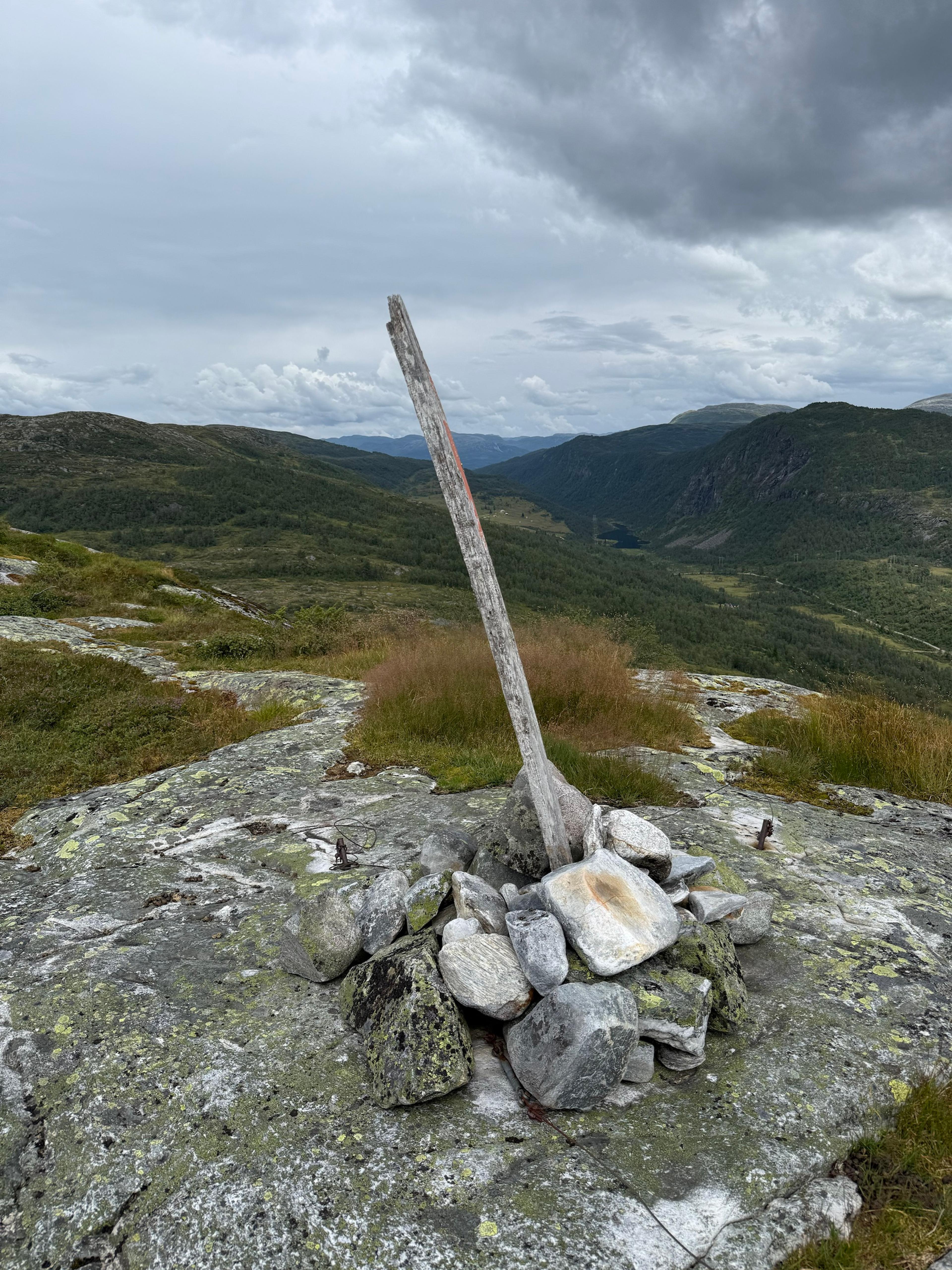 Steinvarde med trestake på bart berg, med en dal og et lite vann nedenfor under mørke skyer.