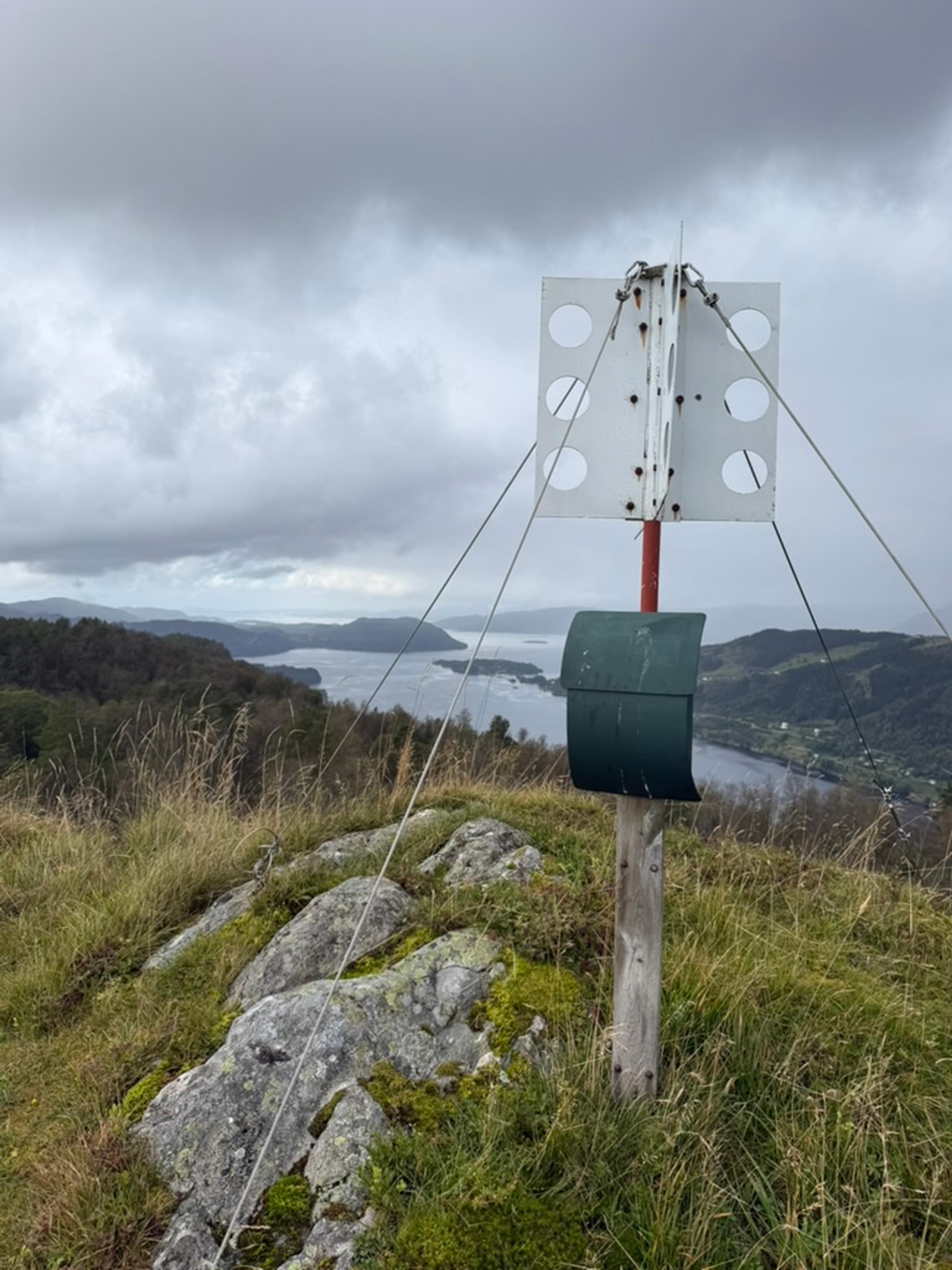 Trigonometrisk punkt med hvite metallplater og en grønn postkasse på en rød og hvit stang, på en gressdekt fjellknaus med lavdekt stein. I bakgrunnen en fjord med holmer og skogkledde åser under grå skyer.