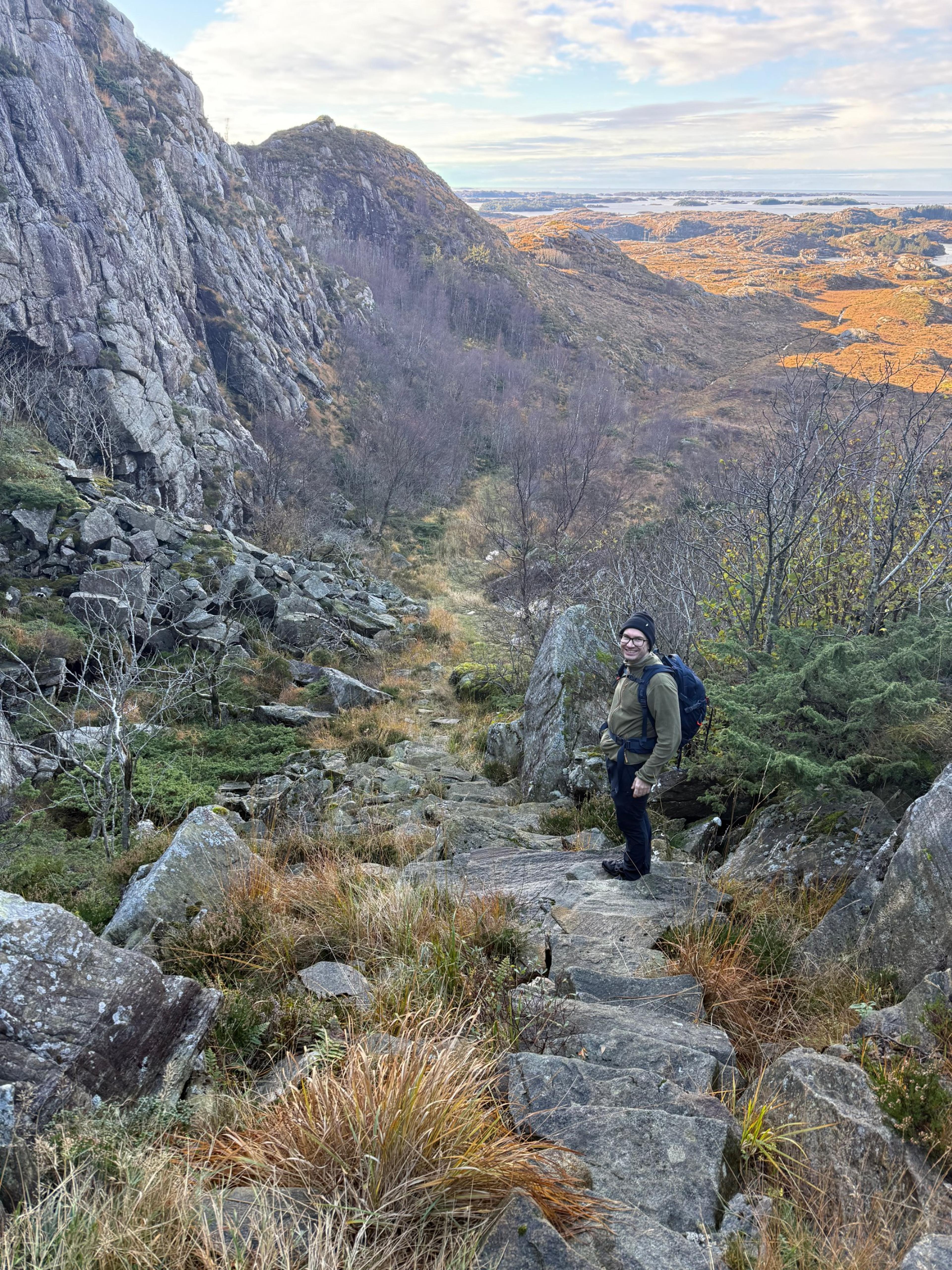 En turgåer med sekk står i en steintrapp som går ned gjennom ur under en høy fjellvegg, med bjørkeskog og lyngmark mot havet nedenfor.
