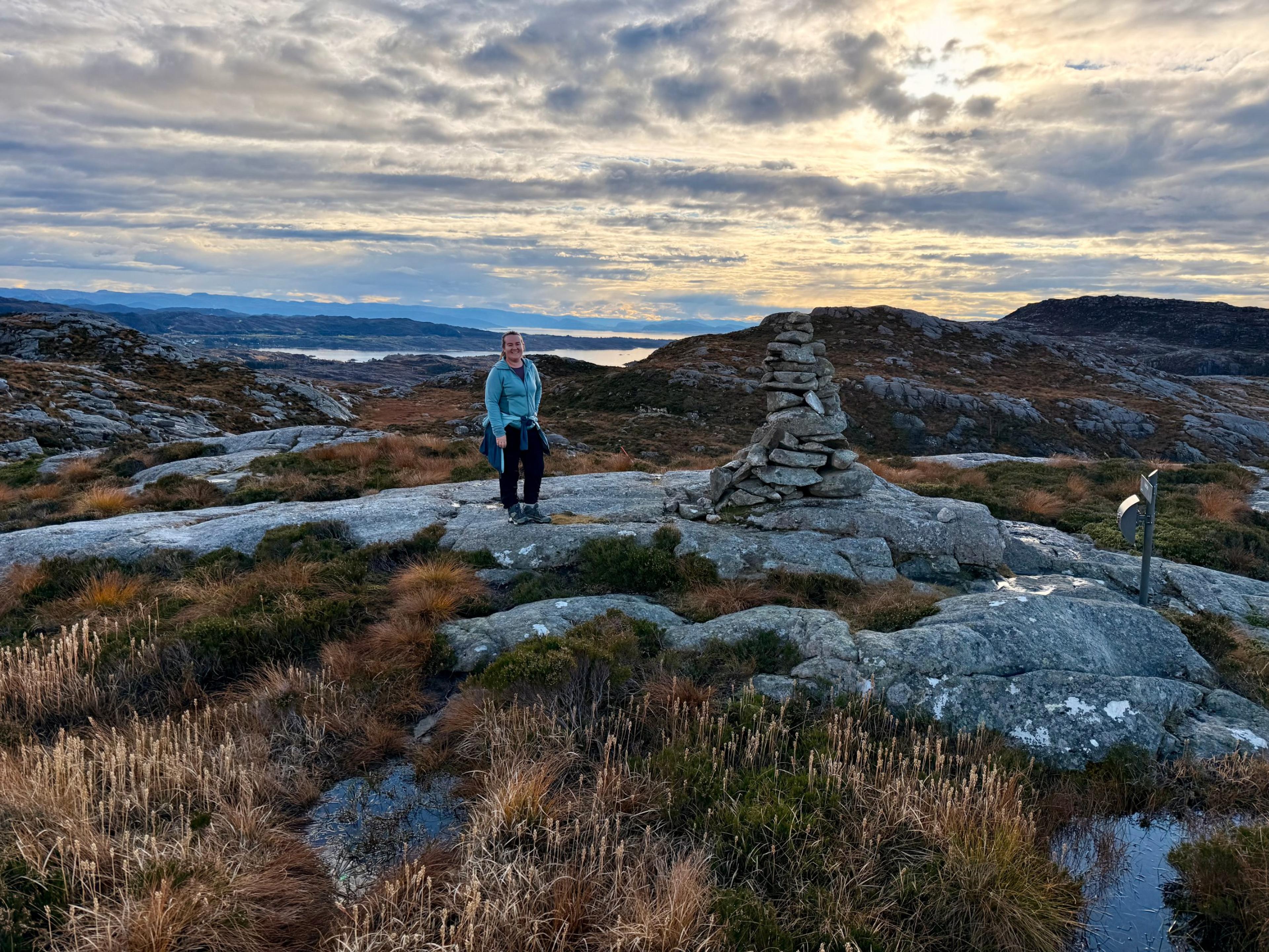 En turgåer står ved en høy steinvarde på et svaberg, med en skiltstolpe til høyre og fjorder og øyer i bakgrunnen under en skydekt himmel.