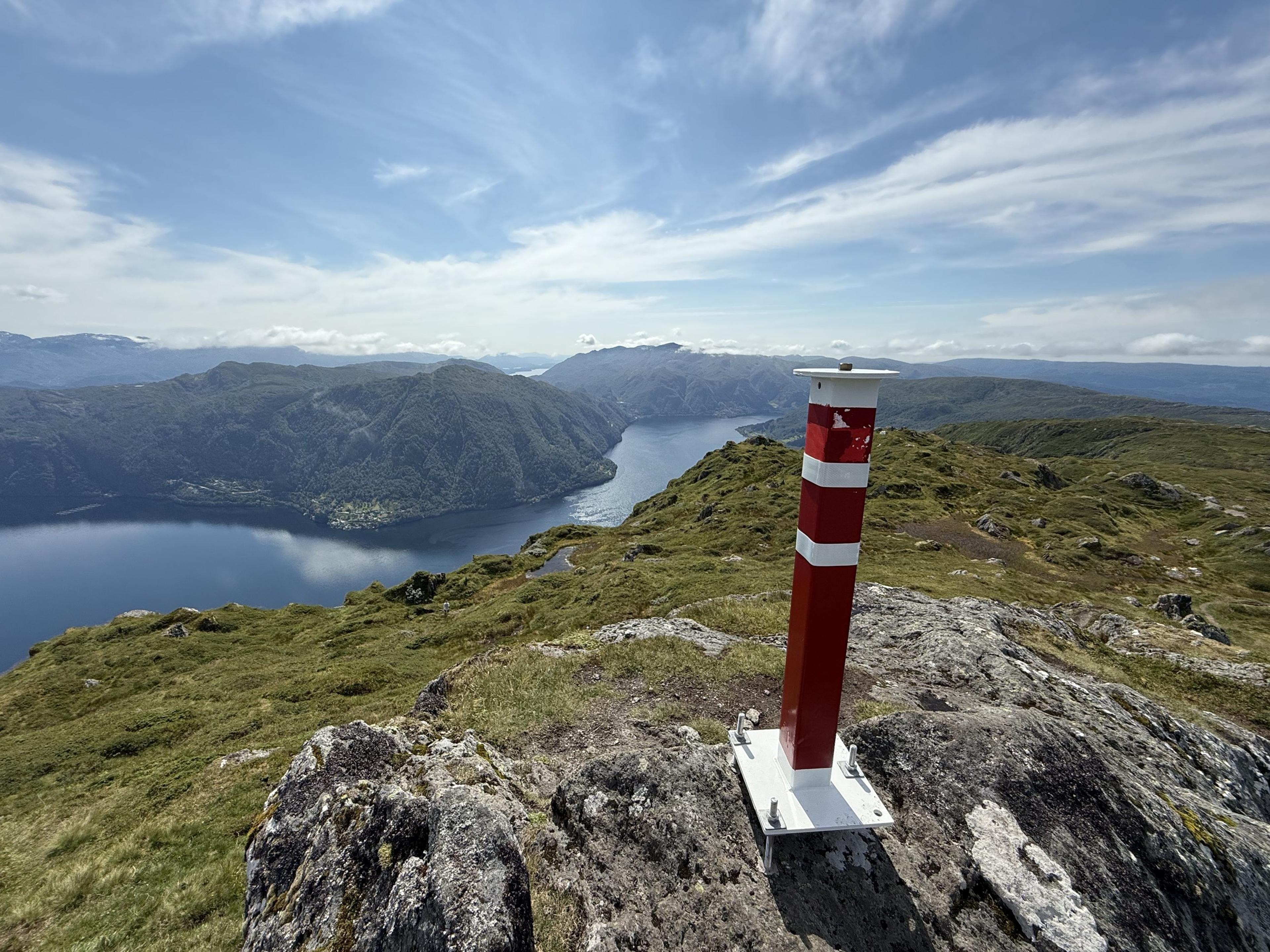Rød og hvit stolpe på en bergknaus, med en fjord mellom bratte, skogkledde fjell og gressdekte rygger rundt.