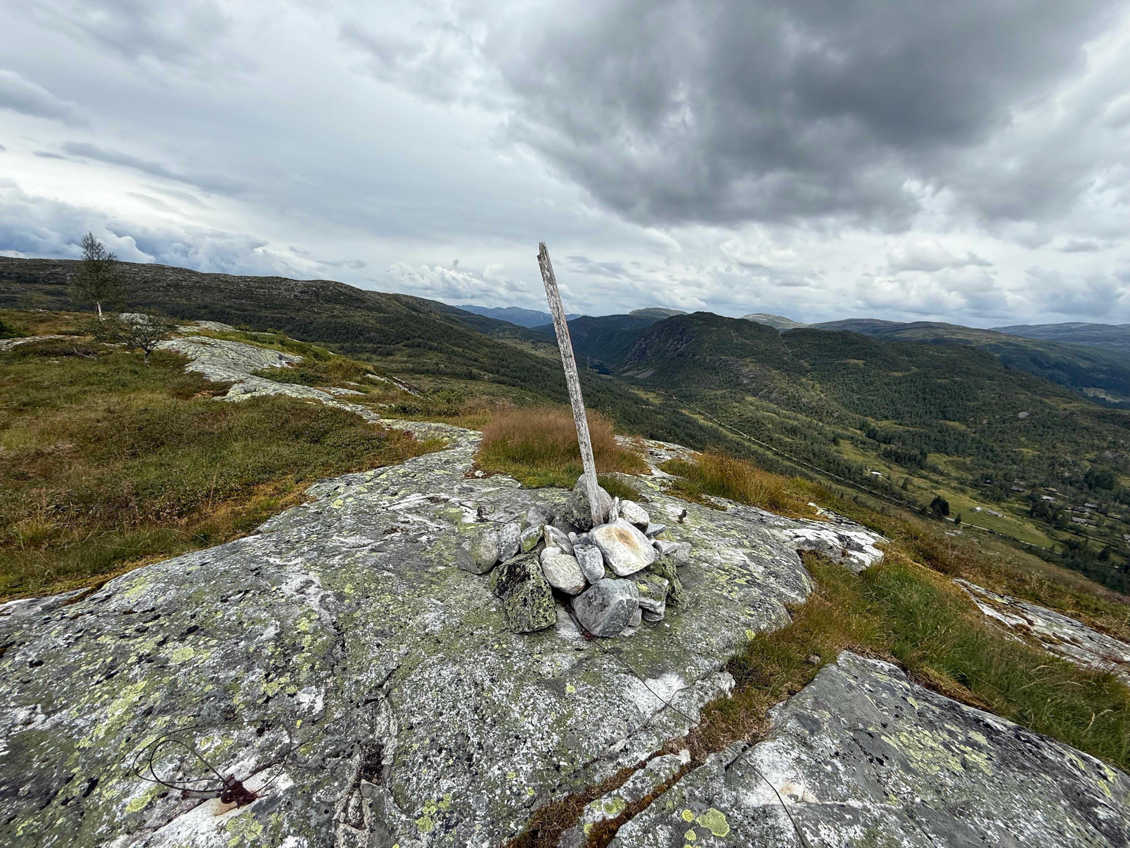 Liten steinvarde med en skjev trestake på lavdekt berg, med åser og en dal med bebyggelse under grå skyer.