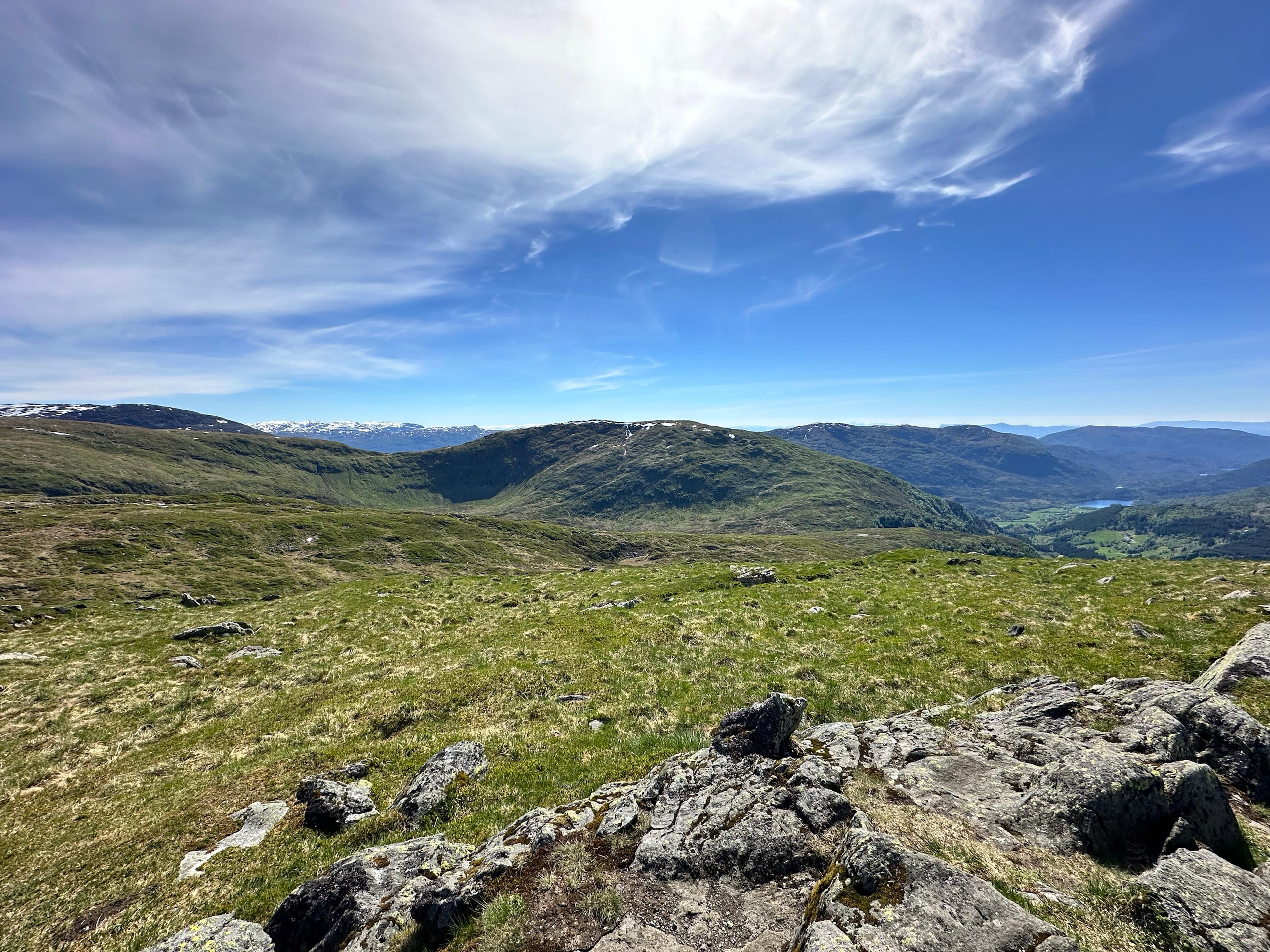 Gresskledd fjellplatå med steiner i forgrunnen, en avrundet fjellrygg i midten og en dal med et lite vann nede til høyre. Snøkledde fjell langt bak.