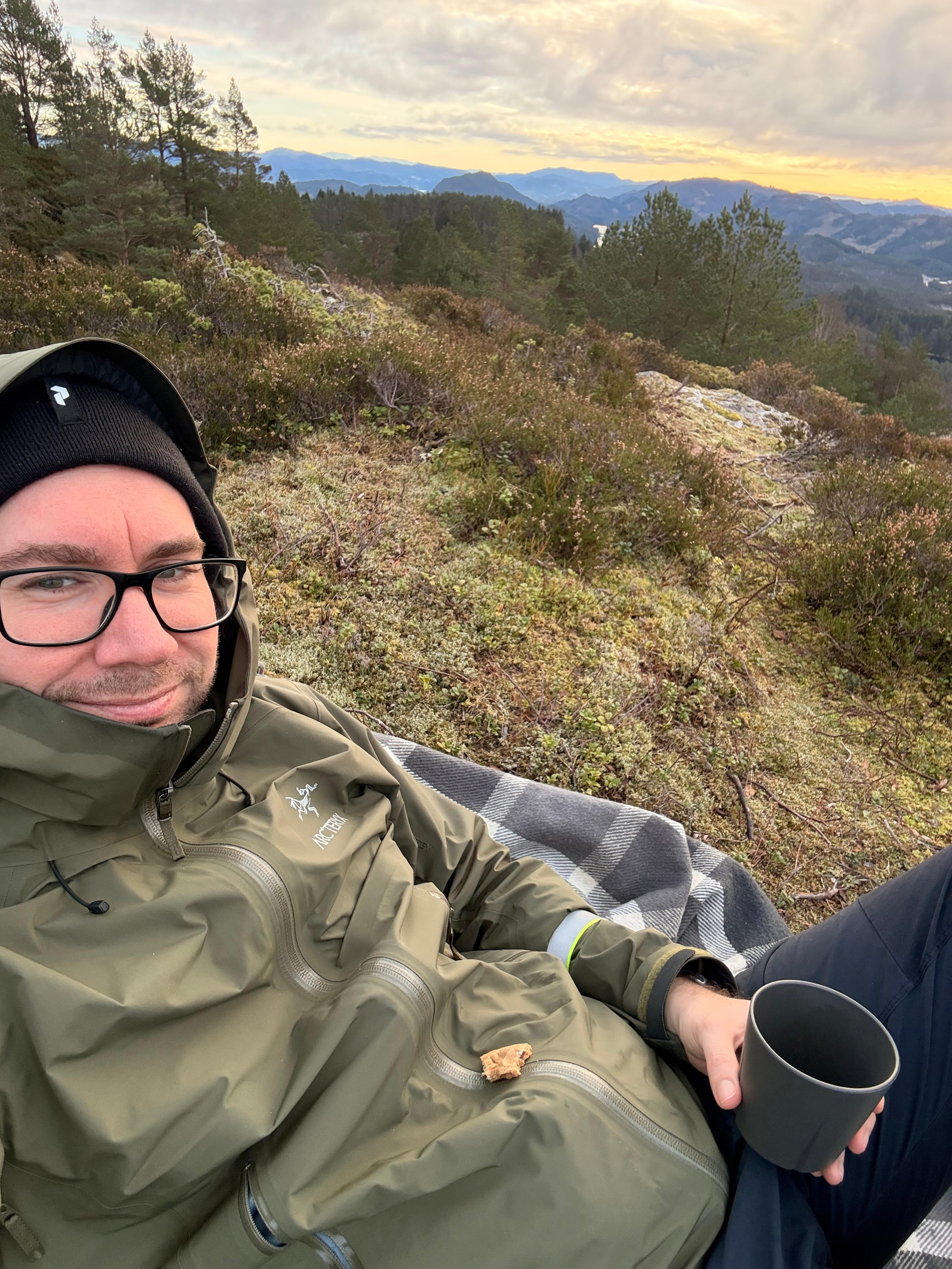 Mann i grønn jakke og lue sitter på et pledd i lyngen med en kaffekopp, med furuskog og fjell bak seg.