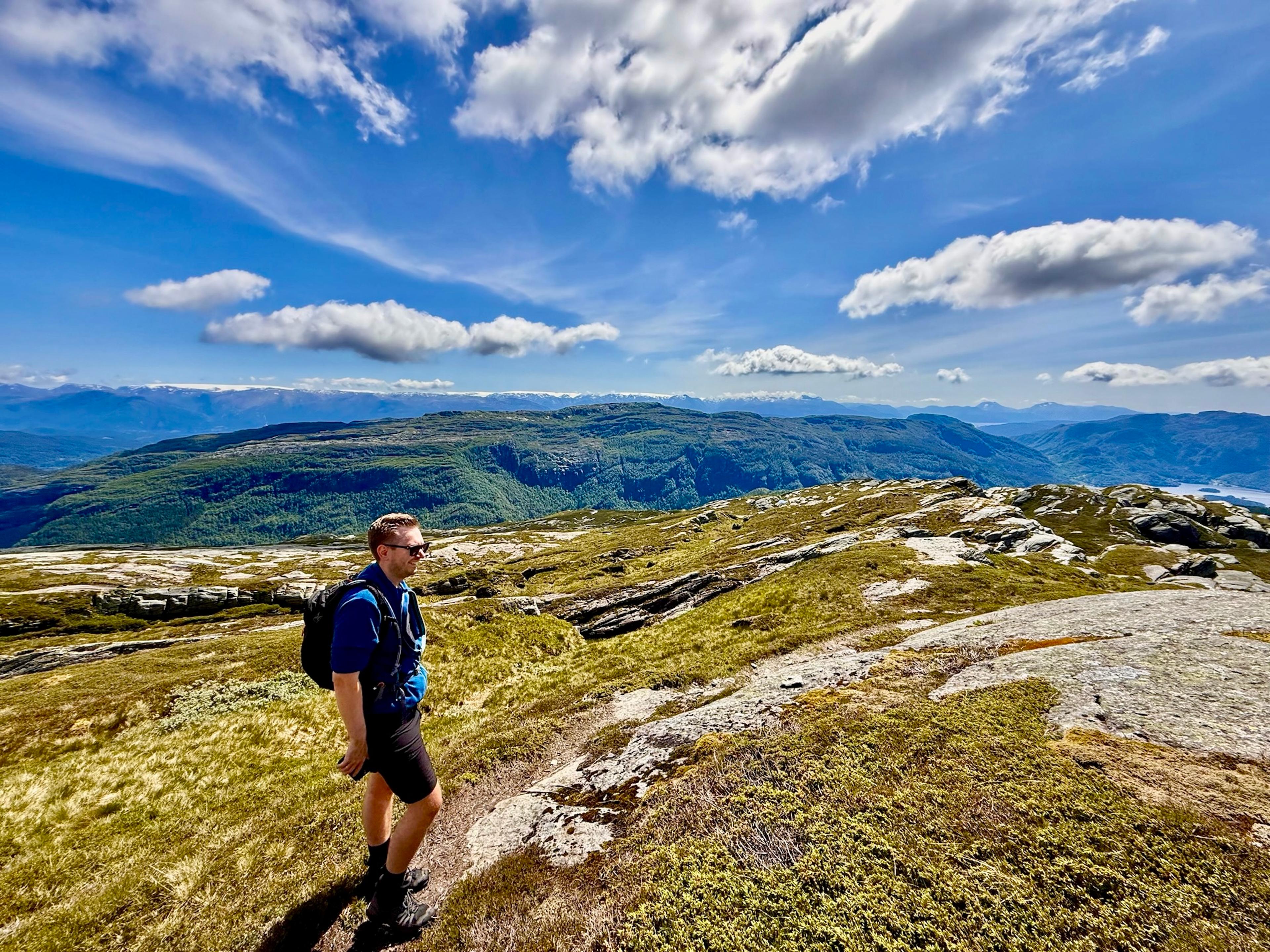 En turgåer i blå genser og shorts står på et svaberg på fjellet. Bak ligger en lang, skogkledd fjellrygg og snødekte fjell langs hele horisonten under blå himmel.