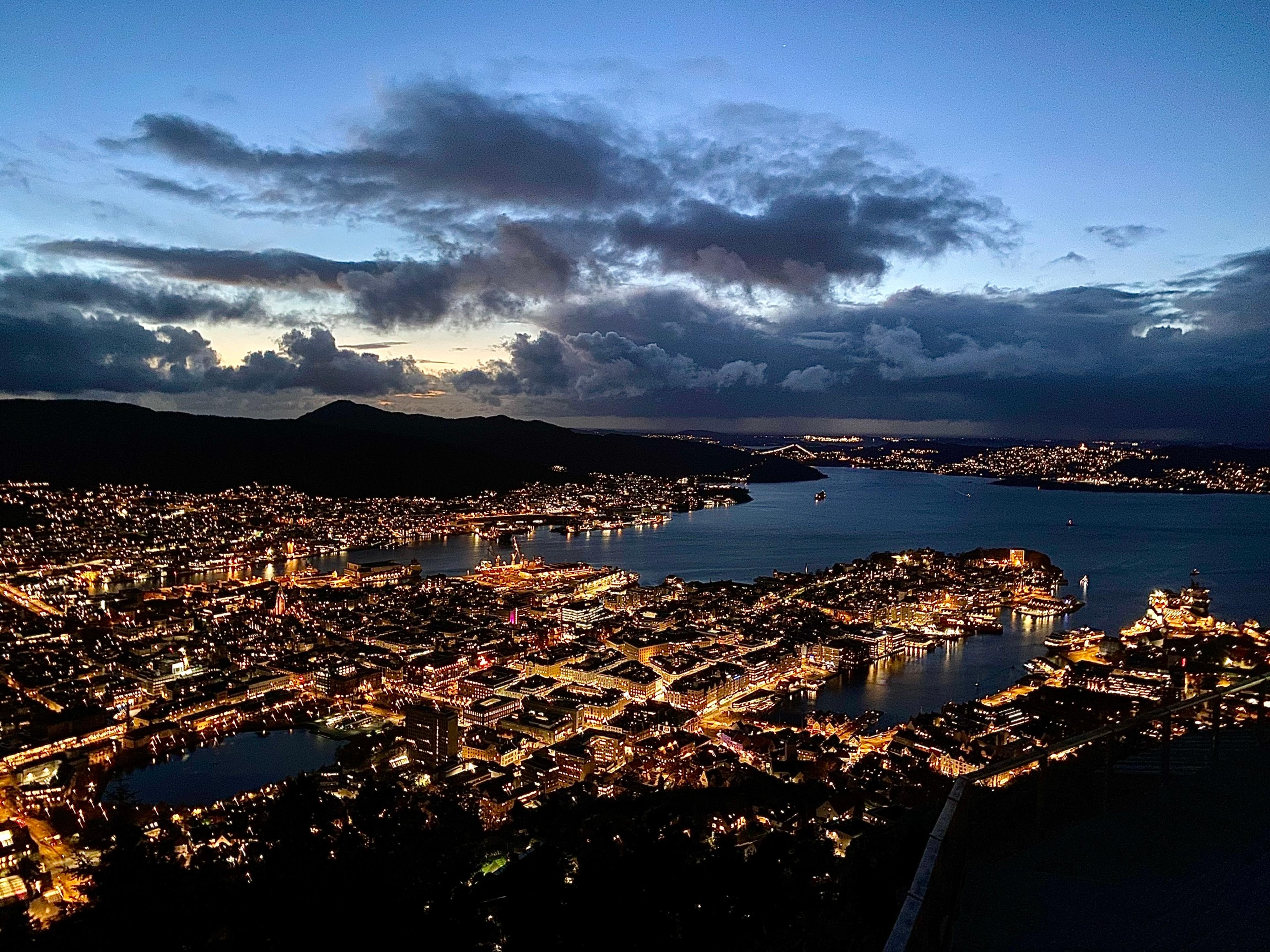 Bergen sentrum i blåtimen sett fra Fløyen. Byen ligger opplyst under et mørkt skydekke, og fjorden strekker seg mot horisonten.